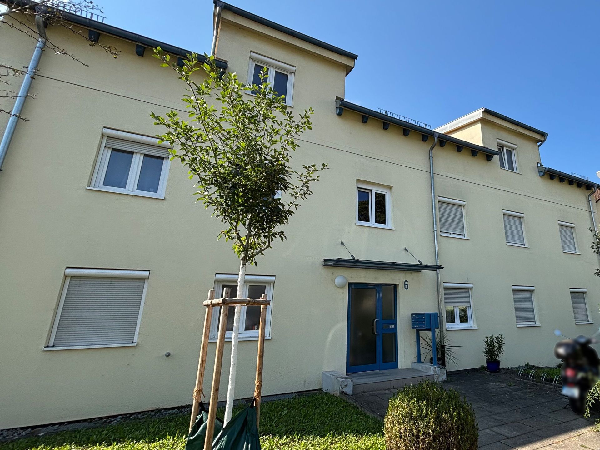 Das Bild zeigt ein Mehrfamilienhaus mit hellgelber Fassade. Vor dem Haus ist ein kleiner Grünstreifen zu sehen mit einem Baum und Sträuchern.
