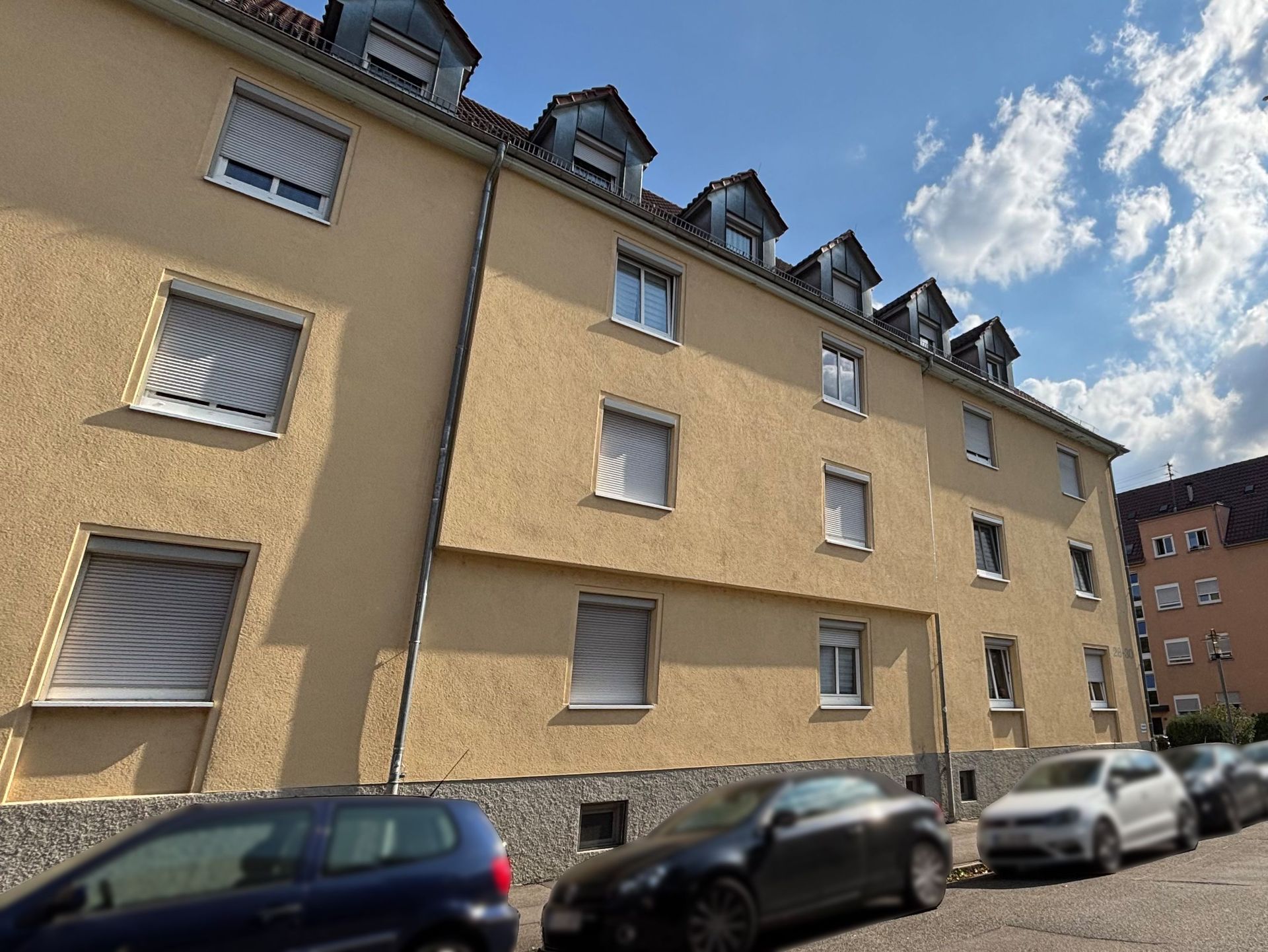 Das Bild zeigt ein Mehrfamilienhaus mit hellgelber Fassade und Dachgauben, vor dem mehrere Autos geparkt sind. Das Gebäude wirkt modern und gepflegt.