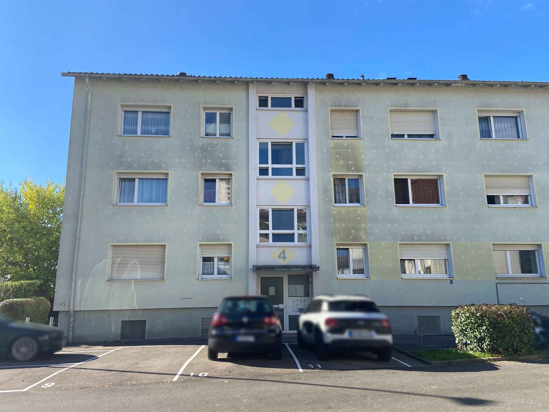 Das Bild zeigt ein Mehrfamilienhaus mit hellgrüner Fassade, das an einigen Stellen Verfärbungen aufweist. Vor dem Haus befinden sich mehrere Parkplätze, auf denen zwei Autos abgestellt sind.