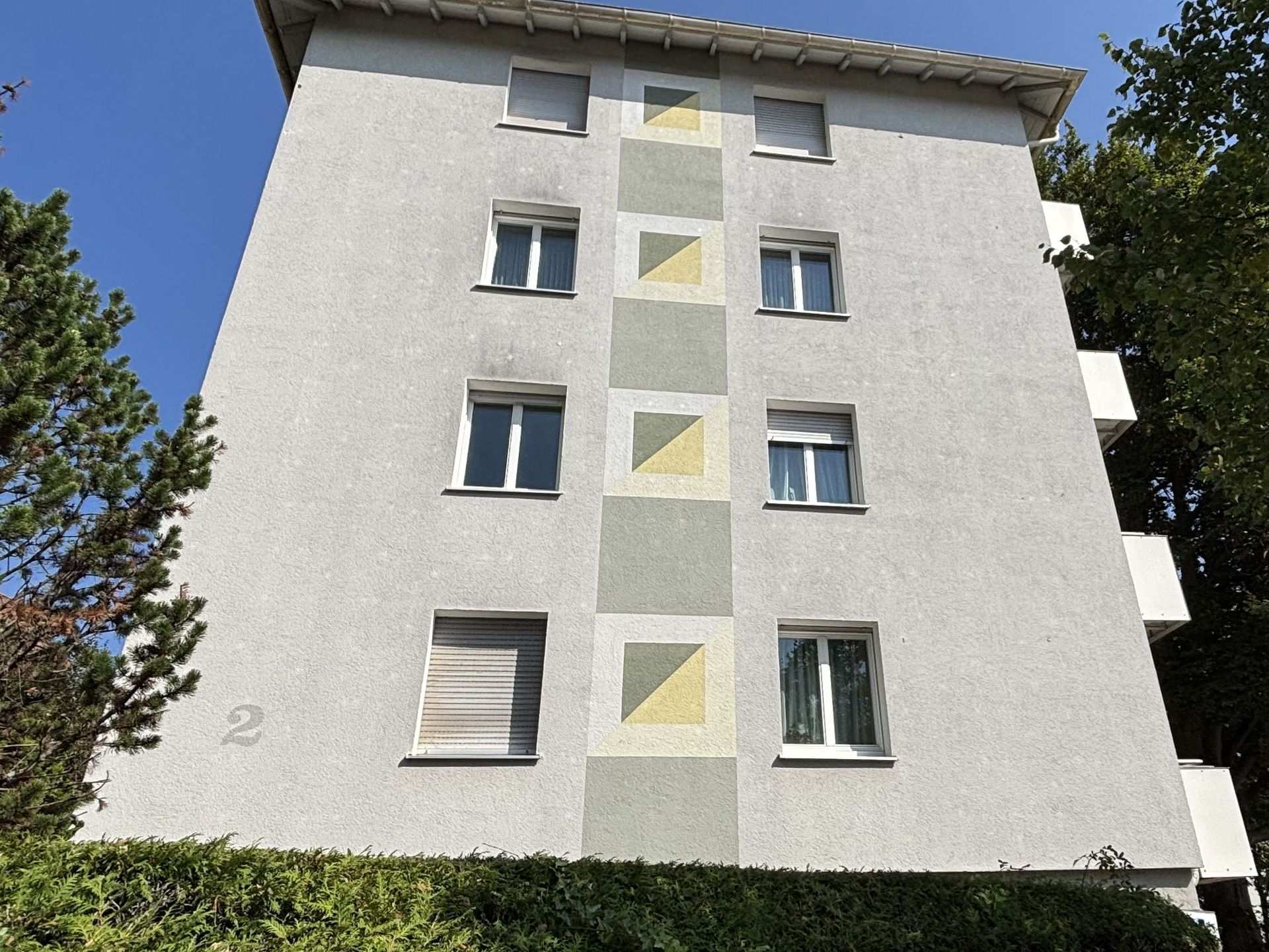 Das Bild zeigt ein Mehrfamilienhaus mit heller Fassade und einer Reihe von gelben und grünen geometrischen Mustern, die zwischen den Fenstern angebracht sind. Vor dem Haus befindet sich eine grüne Hecke, die den unteren Bereich verdeckt.