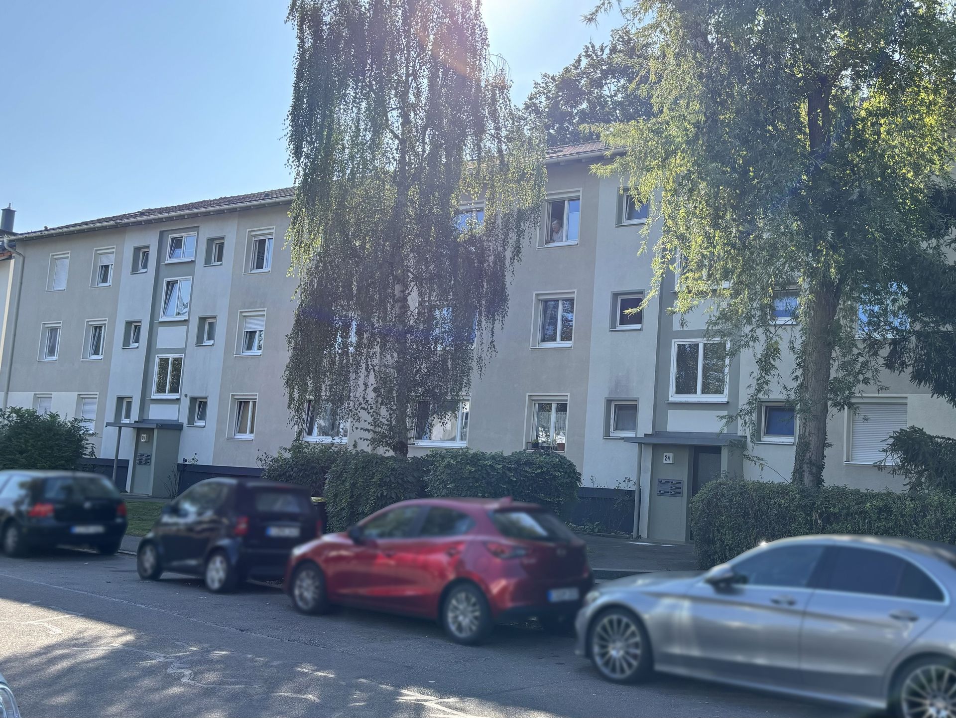 Das Bild zeigt ein Mehrfamilienhaus mit heller Fassade und vielen Fenstern. Vor dem Haus stehen mehrere Autos am Straßenrand.
