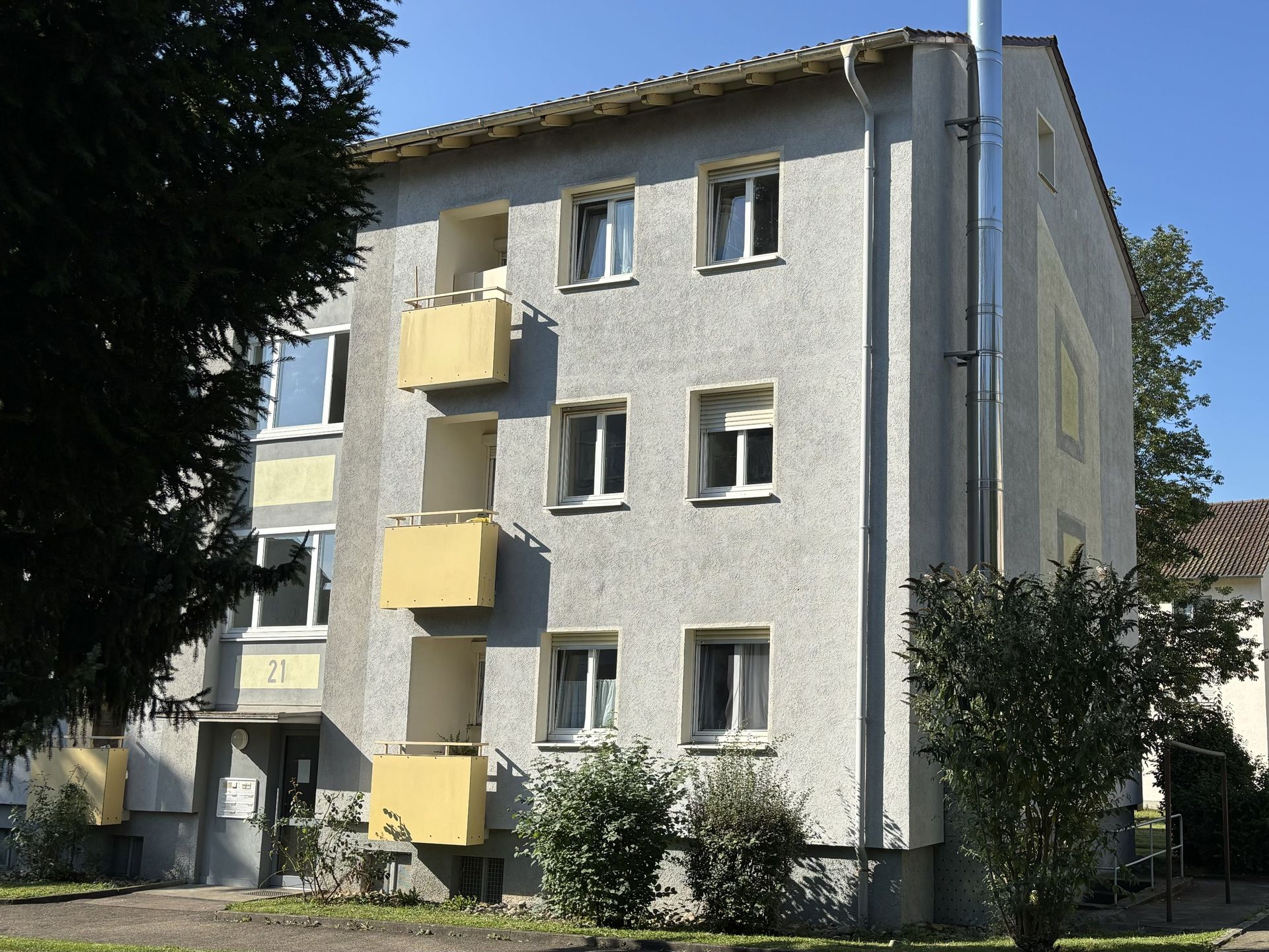 Das Bild zeigt ein Mehrfamilienhaus mit grauer Fassade und gelben Balkonen. Neben dem Haus steht ein Baum, und im Hintergrund ist ein weiteres Gebäude zu sehen.