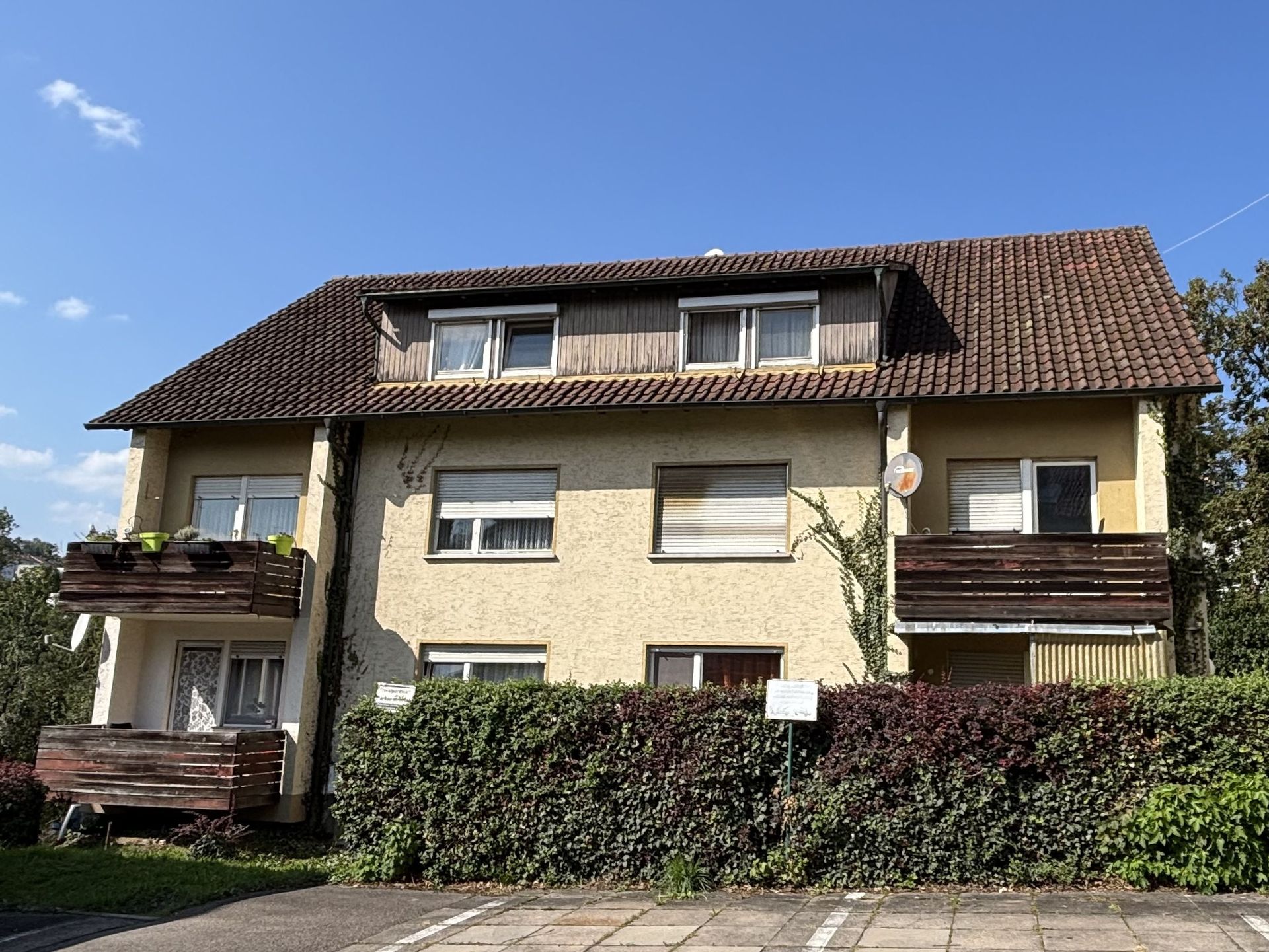 Das Bild zeigt ein Mehrfamilienhaus mit hellgelber Fassade, braunem Dach und mehreren Balkonen.  Vor dem Haus befindet sich eine dichte Hecke, die das Erdgeschoss fast vollständig verdeckt.