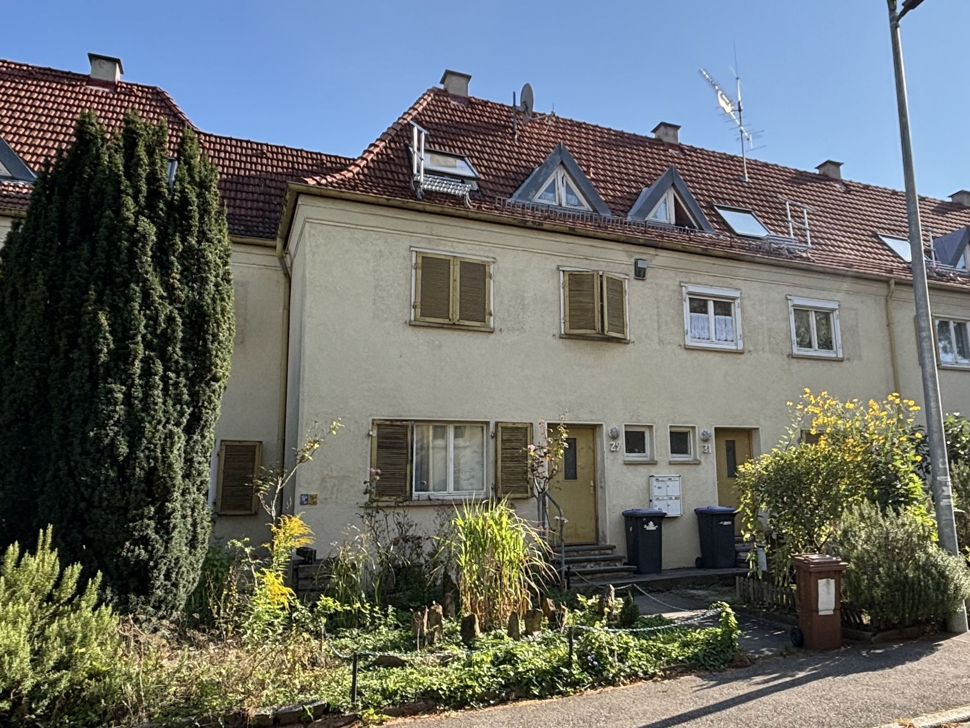 Das Bild zeigt eine Reihe von Häusern mit heller Fassade, roten Ziegeldächern und gelben Fensterläden. Vor den Häusern befinden sich kleine Vorgärten mit unterschiedlicher Bepflanzung.