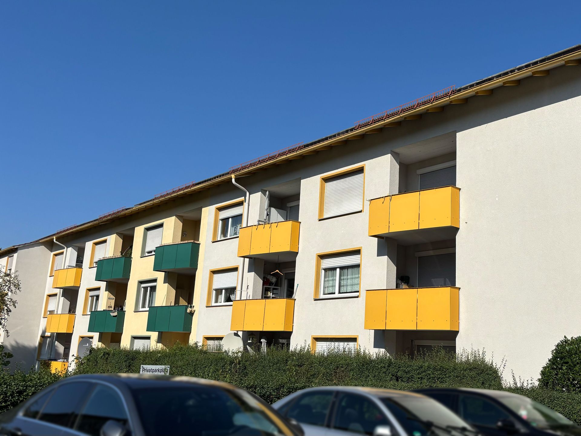 Das Bild zeigt ein Mehrfamilienhaus mit hellgelber Fassade und dunkelblauen Akzenten um die Fenster.  Vor dem Haus befinden sich Sträucher und ein kleiner Baum.