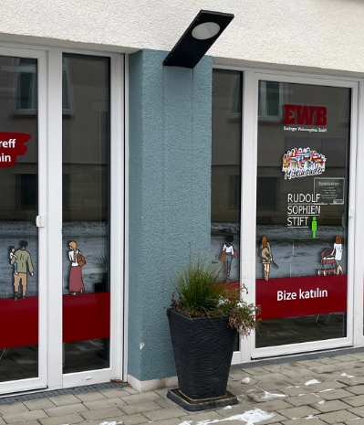 Das Bild zeigt einen Eingangsbereich eines Gebäudes. Die Fenster sind mit Gestaltungselementen beklebt und eine Pflanze steht dort. Link zum Bereich Nachbarschaftstreff Am Schönen Rain.