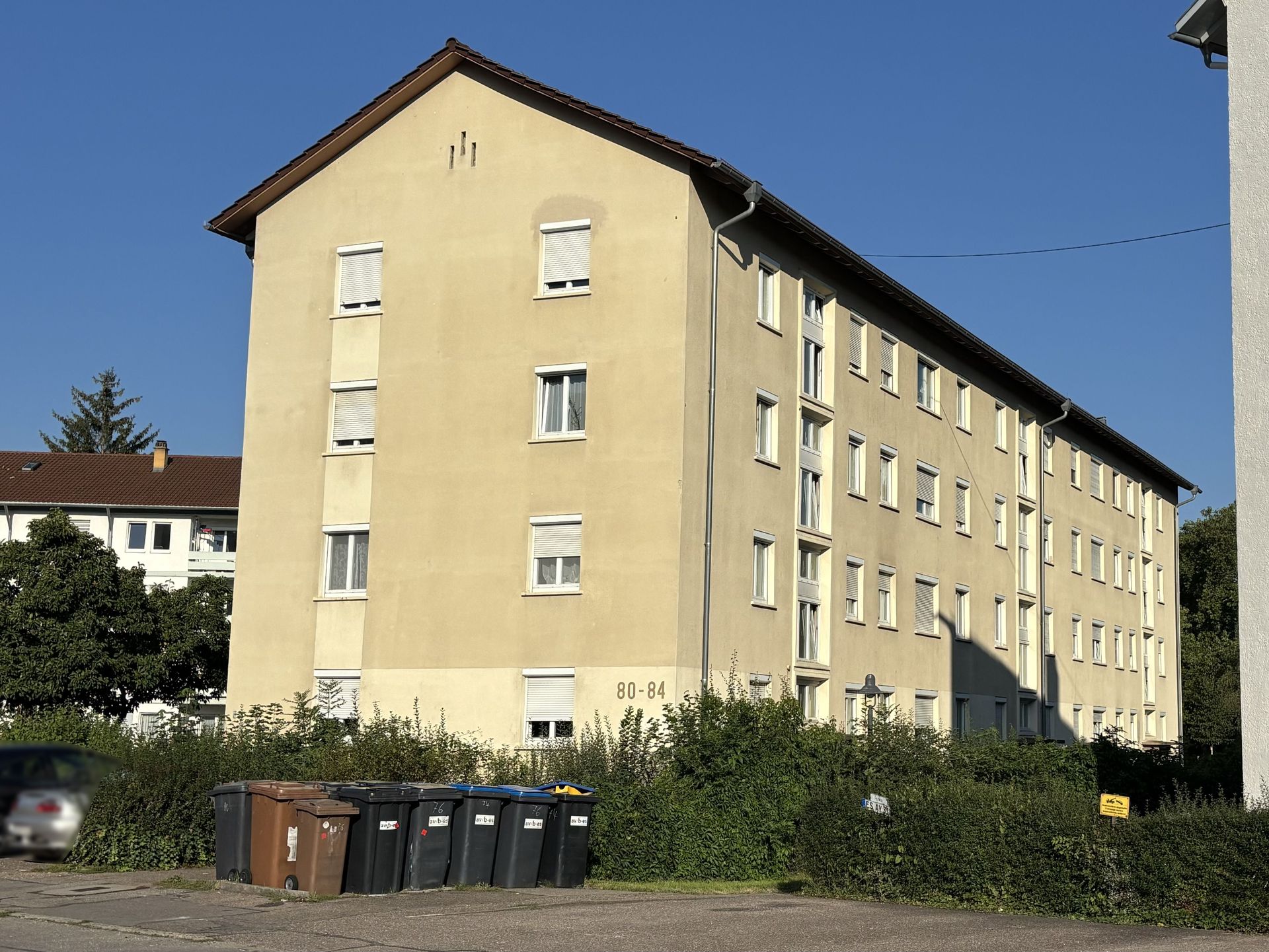 Das Bild zeigt ein Mehrfamilienhaus mit hellgelber Fassade und vielen Fenstern. Vor dem Haus stehen Mülltonnen und eine grüne Hecke.