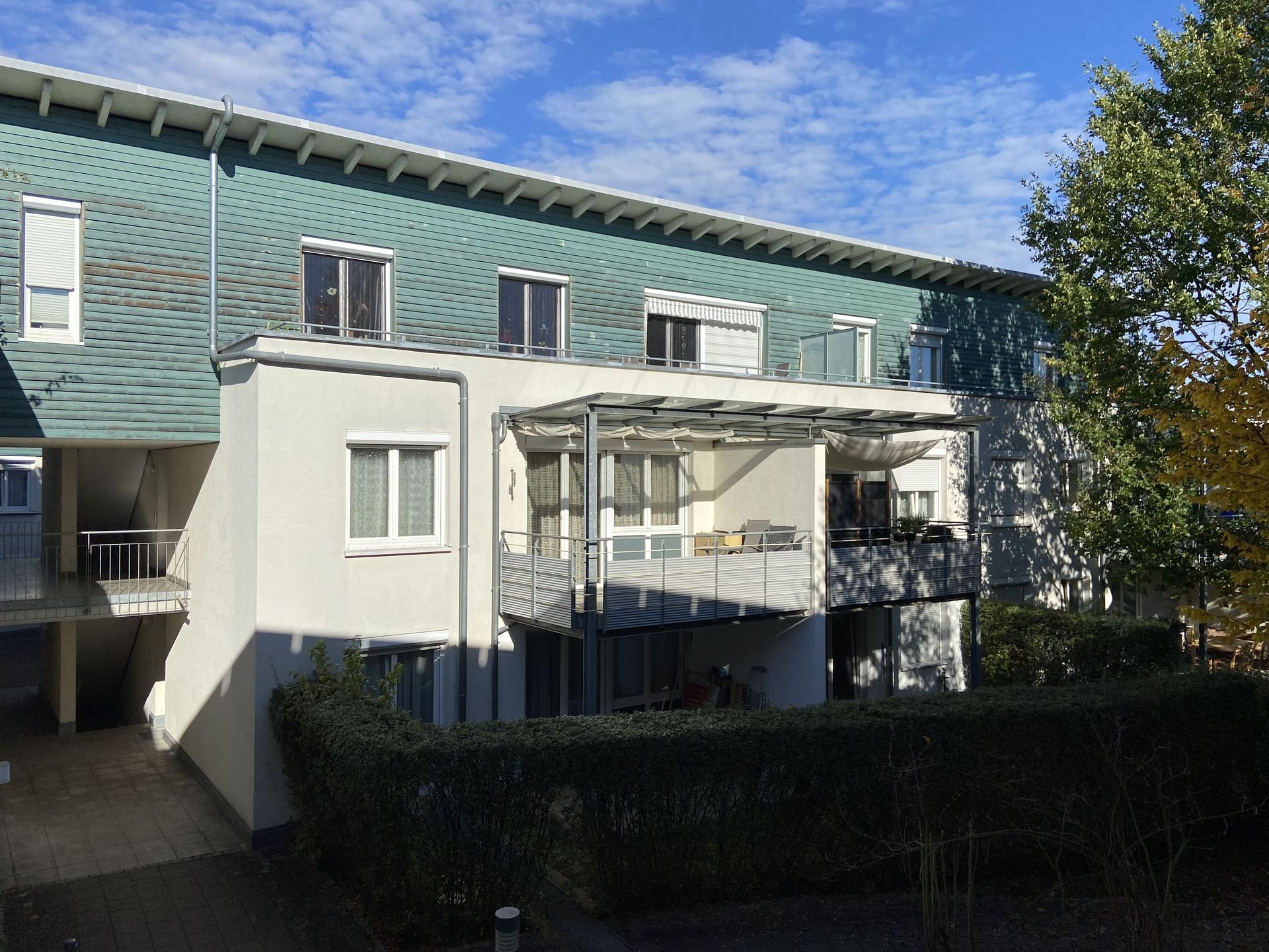 Das Bild zeigt ein modernes Mehrfamilienhaus mit weißer Fassade und einem auffälligen, türkisfarbenen Holzelement im oberen Bereich. Vor dem Haus befindet sich eine Terrasse mit Überdachung und eine grüne Hecke.