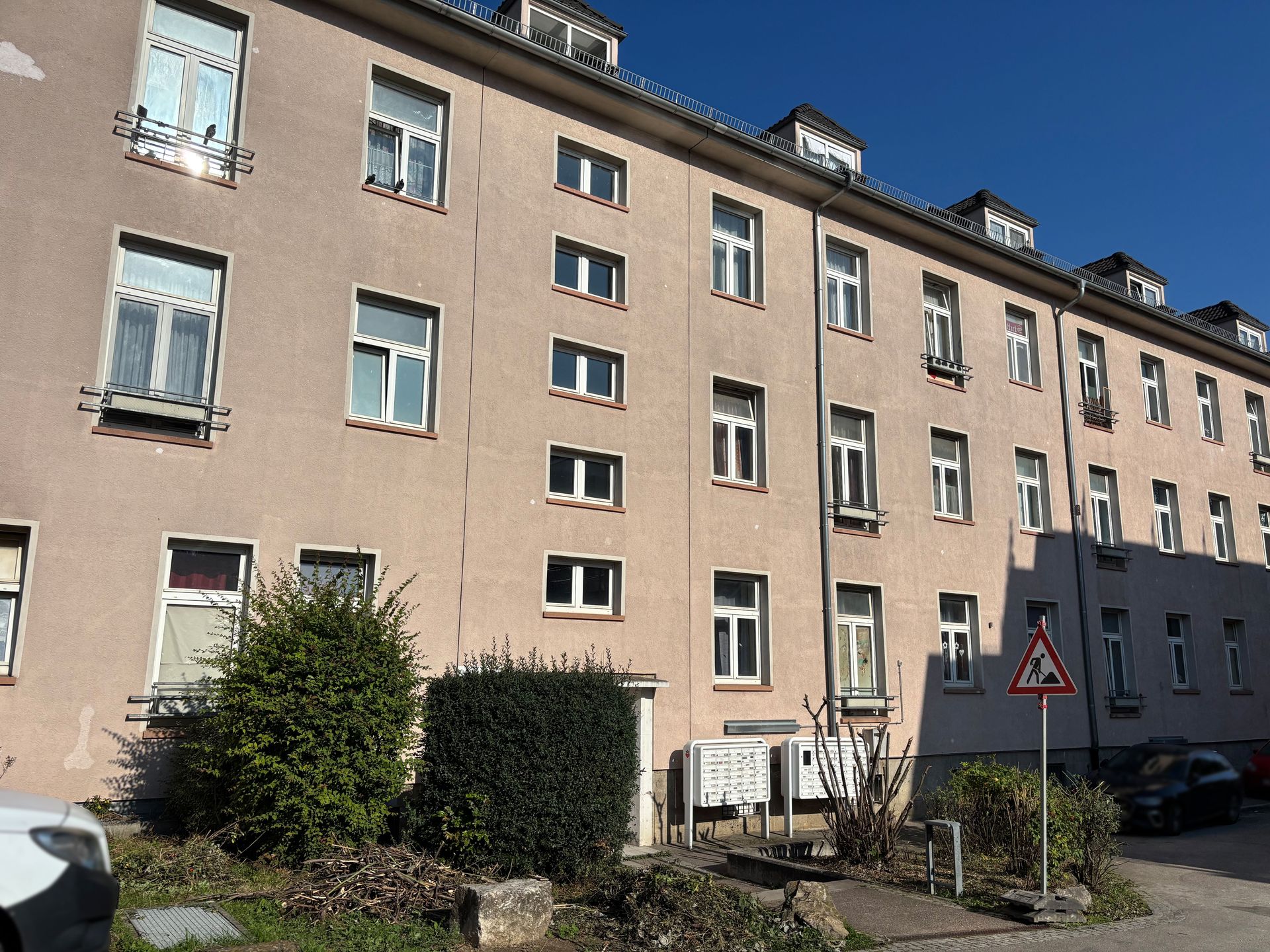 Das Bild zeigt ein helles Mehrfamilienhaus mit vielen Fenstern. Vor dem Haus befindet sich ein gepflasterter Bereich mit Briefkästen und grünen Büschen.
