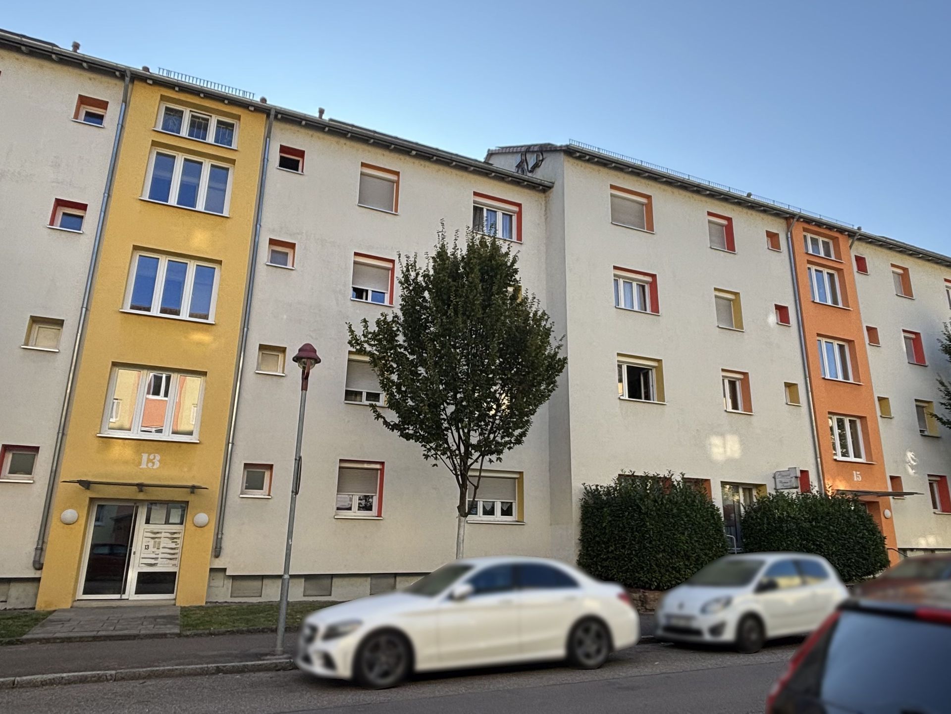 Das Bild zeigt ein Mehrfamilienhaus mit heller Fassade und farbigen Akzenten an den Fenstern und Eingängen. Vor dem Haus stehen zwei Autos am Straßenrand und ein Baum.