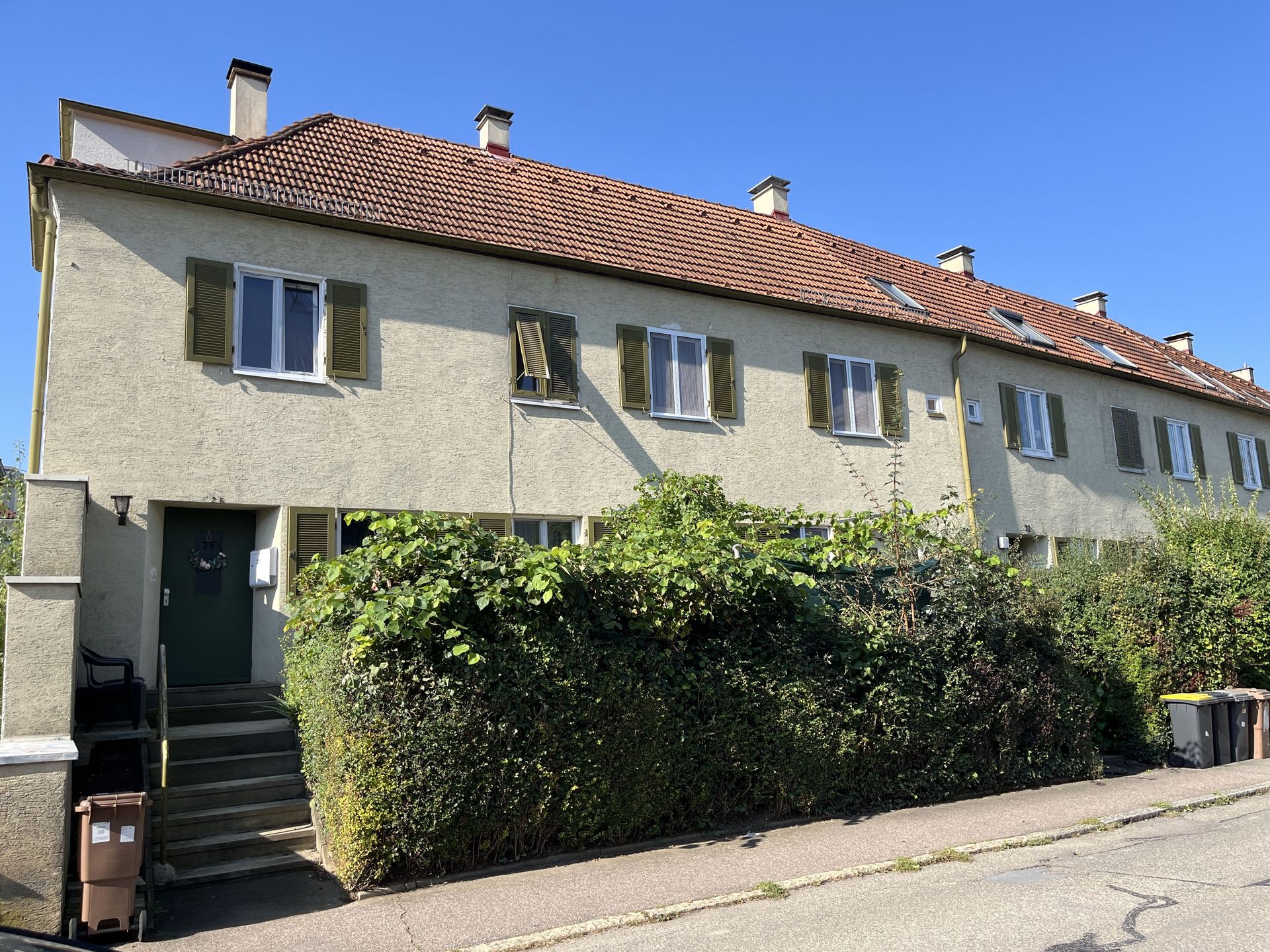 Das Bild zeigt eine Reihe von Häusern mit heller Fassade, roten Ziegeldächern und grünen Fensterläden.  Vor den Häusern befindet sich eine dichte Hecke, die die Sicht auf die unteren Stockwerke verdeckt.