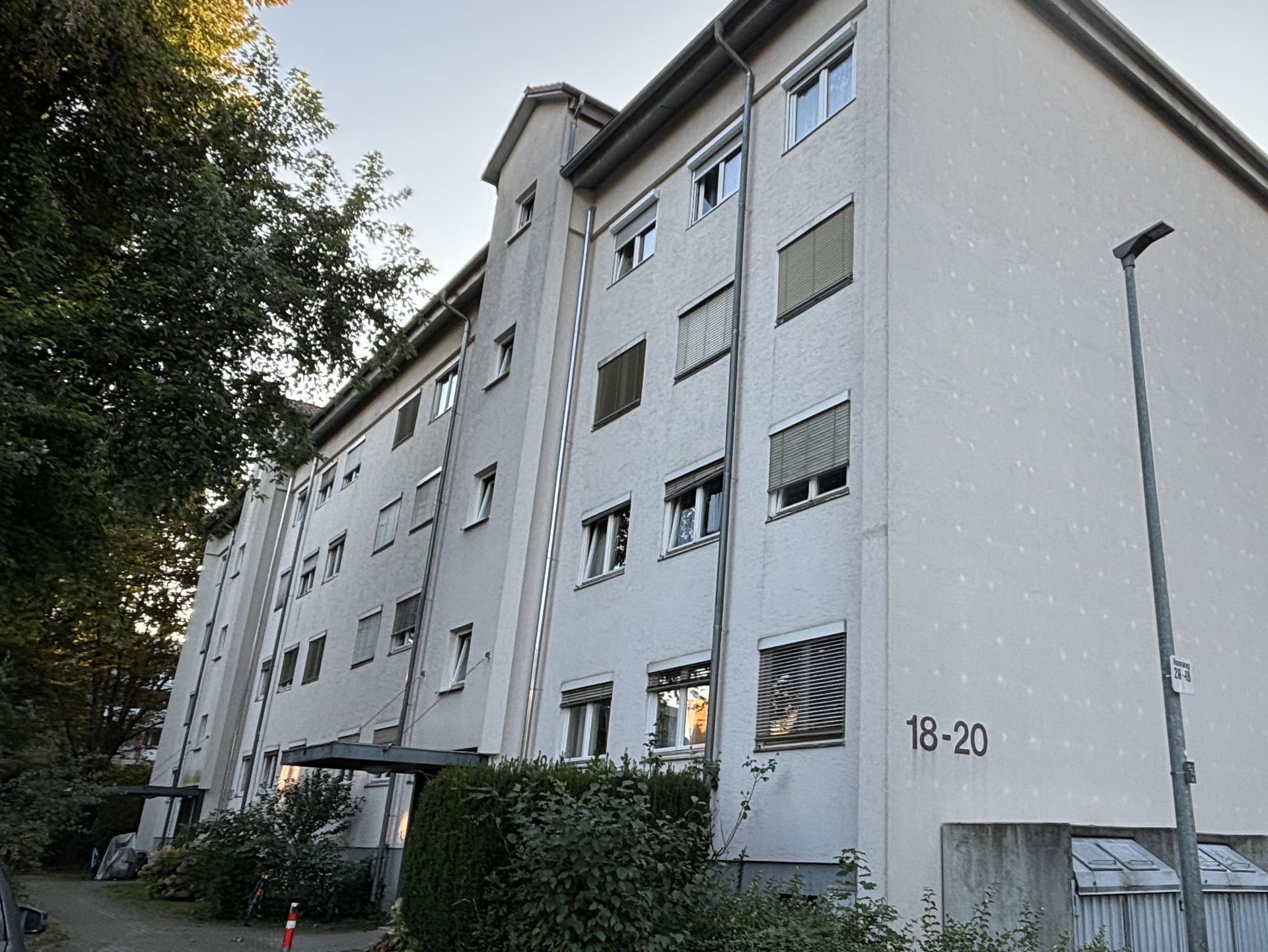 Das Bild zeigt ein Mehrfamilienhaus mit heller Fassade. Vor dem Haus befindet sich ein kleiner gepflasterter Bereich mit Hecke und einem Baum.