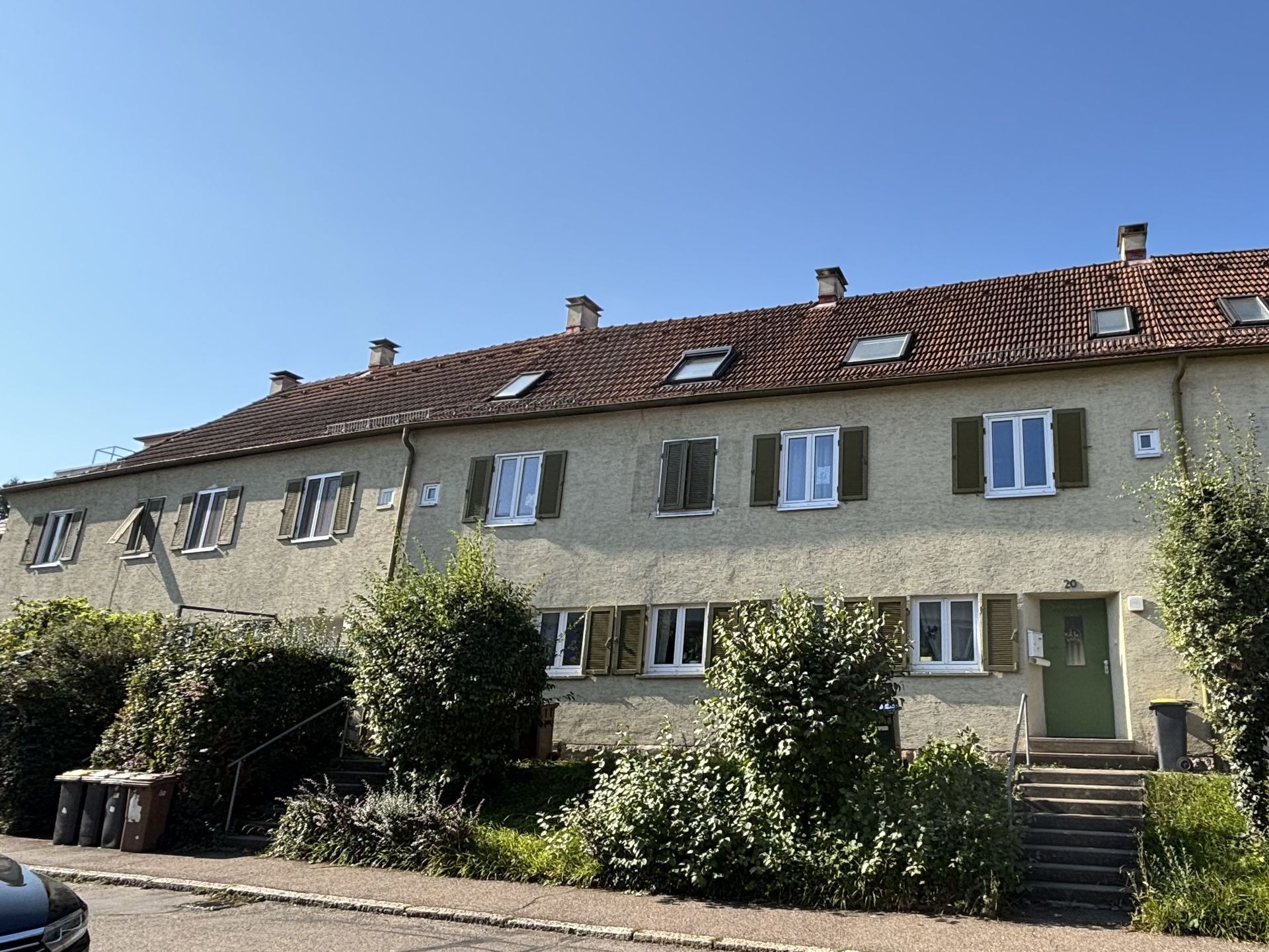 Das Bild zeigt eine Reihe von Häusern mit heller Fassade, roten Ziegeldächern und grünen Fensterläden. Vor den Häusern befinden sich kleine Vorgärten mit Sträuchern und eine Treppe führt zum Eingang der Häuser.