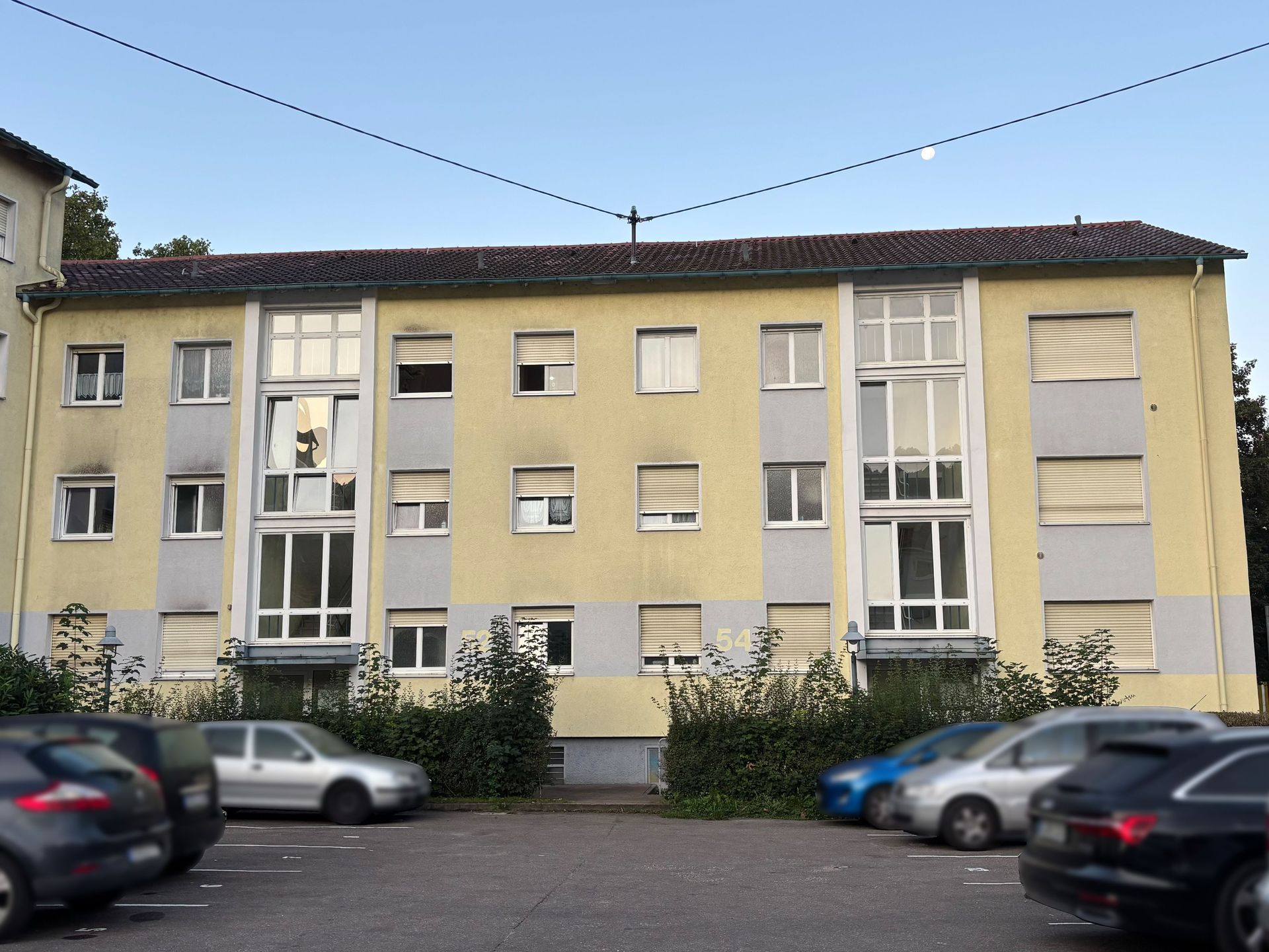 Das Bild zeigt ein Mehrfamilienhaus mit hellgelber Fassade und grauen Elementen, vor dem mehrere Autos auf einem Parkplatz abgestellt sind. Das Gebäude hat mehrere Stockwerke mit gleichmäßig angeordneten Fenstern.