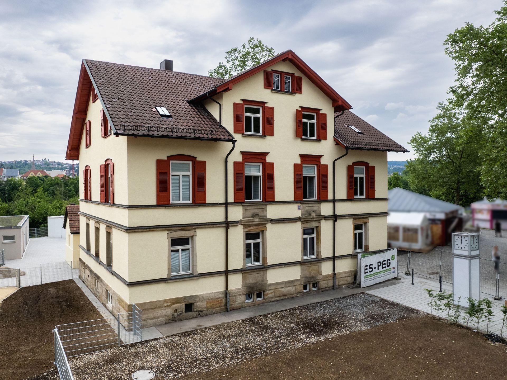 Das Bild zeigt ein helles Gebäude mit braunem Dach und braunen Fensterläden. Vor dem Haus befindet sich eine unbefestigte Fläche und im Hintergrund sind Bäume zu sehen.
