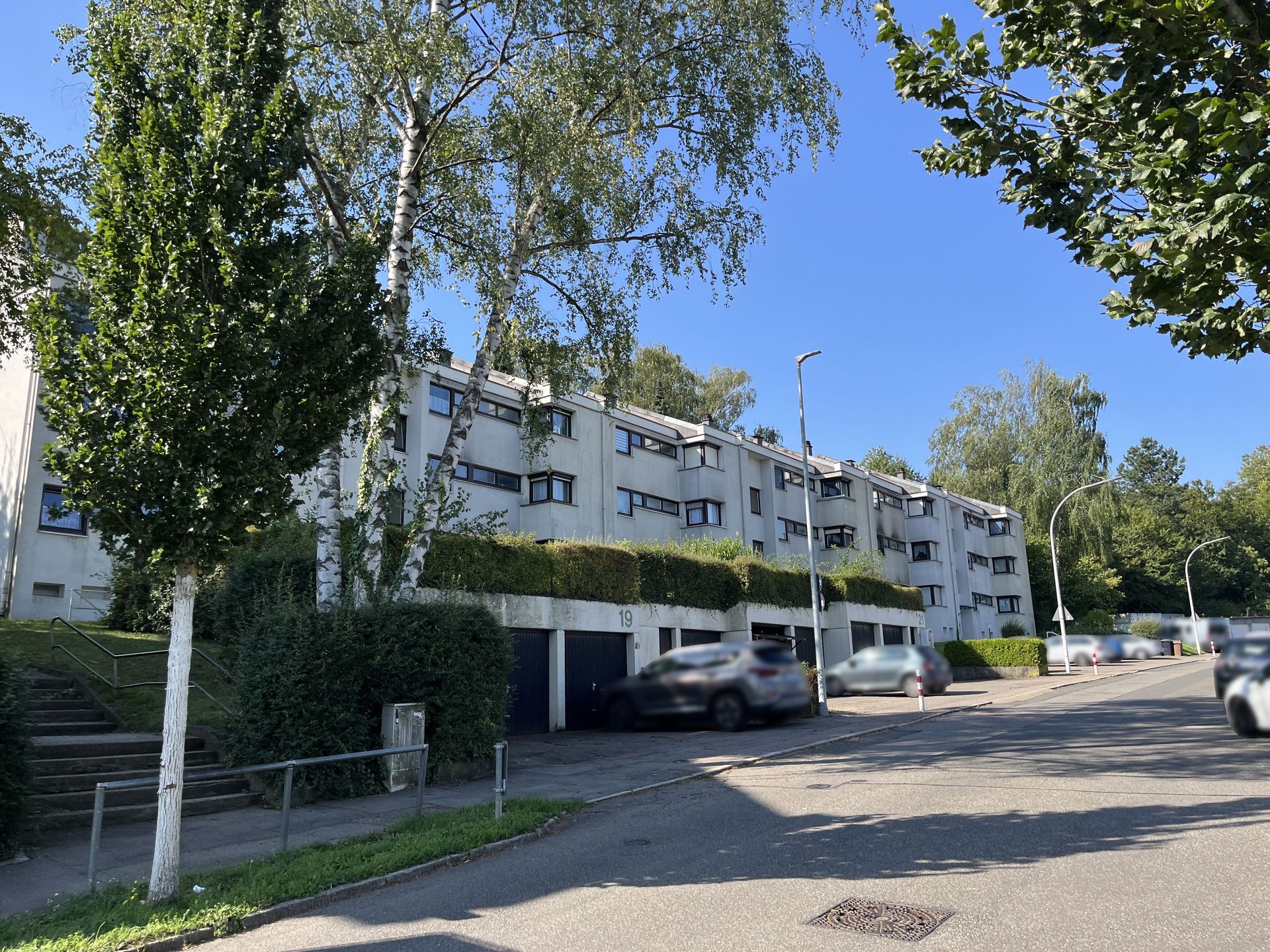 Das Bild zeigt ein langgezogenes Mehrfamilienhaus mit heller Fassade und mehreren Eingängen. Vor dem Haus befinden sich Garagen und einige Bäume säumen die Straße.