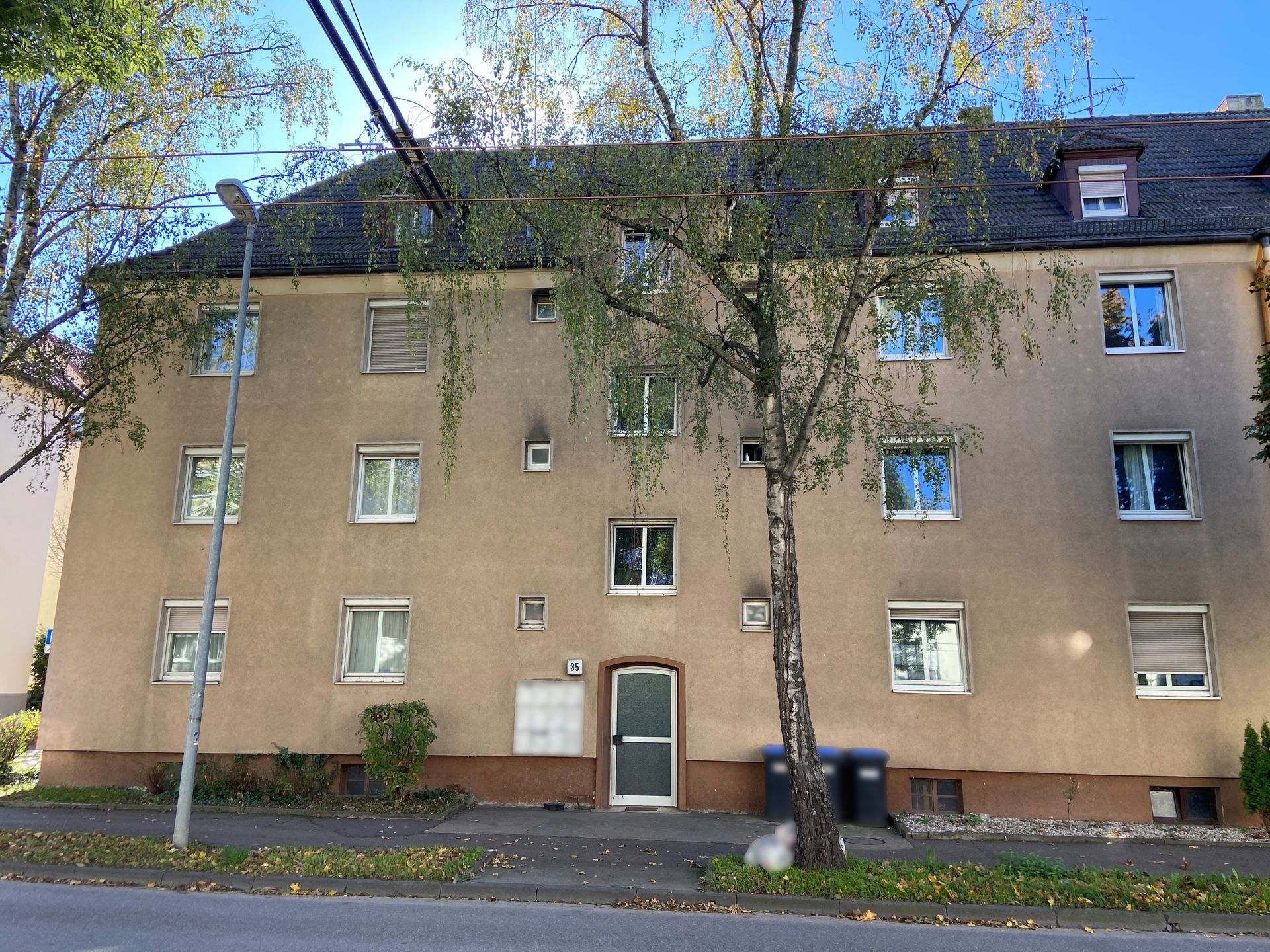 Das Bild zeigt ein Mehrfamilienhaus mit heller Fassade und dunklem Dach. Vor dem Haus befindet sich ein Baum und am Straßenrand stehen Mülltonnen.