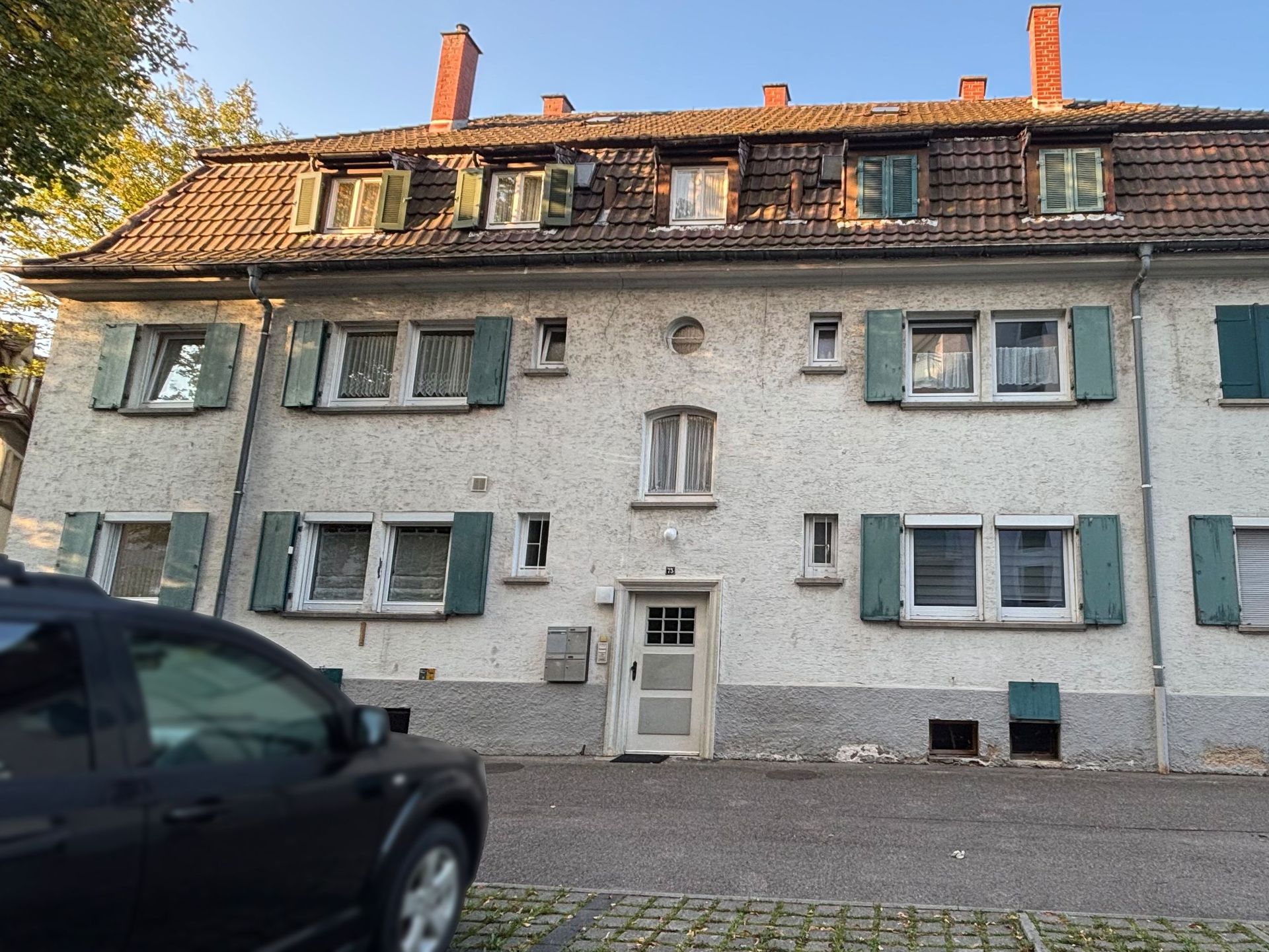 Das Bild zeigt ein mehrstöckiges Mehrfamilienhaus mit weißer Fassade und grünen Fensterläden. Vor dem Haus befindet sich eine gepflasterte Straße und ein geparktes Auto.