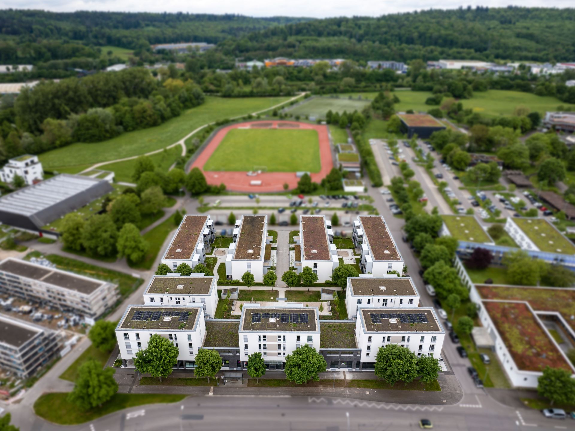 Das Bild zeigt eine Wohnanlage mit mehreren Häusern. Im Hintergrund sieht man einen Sportplatz und viele große Grünflächen sowie Bäume.