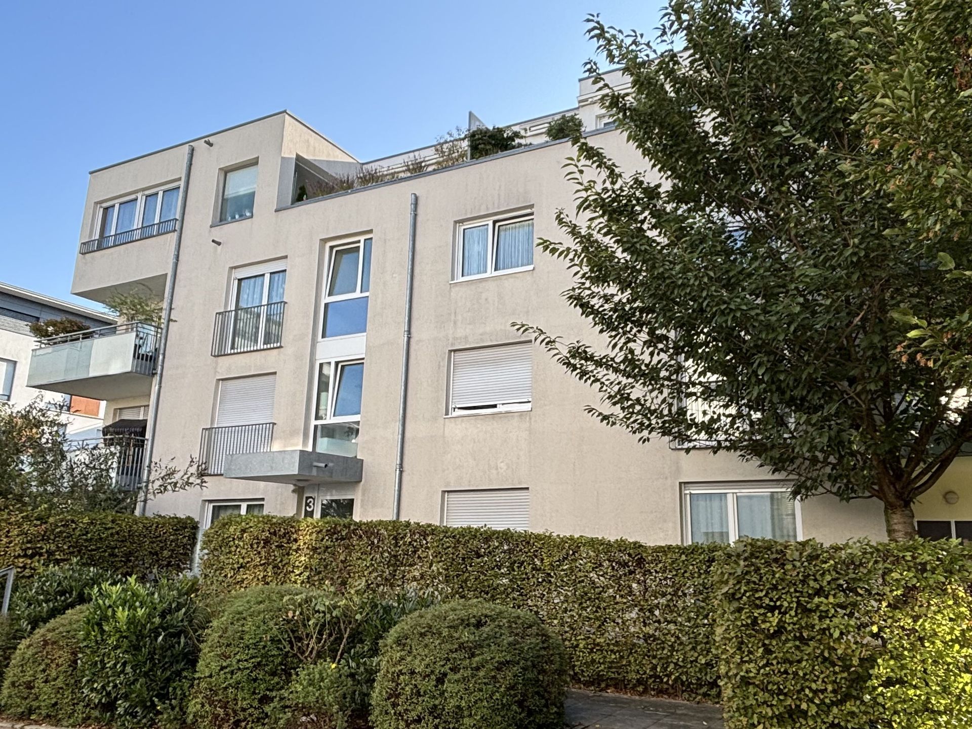 Das Bild zeigt ein modernes Mehrfamilienhaus mit heller Fassade und Balkonen. Vor dem Haus befindet sich eine hohe, dichte Hecke, die das Erdgeschoss verdeckt.