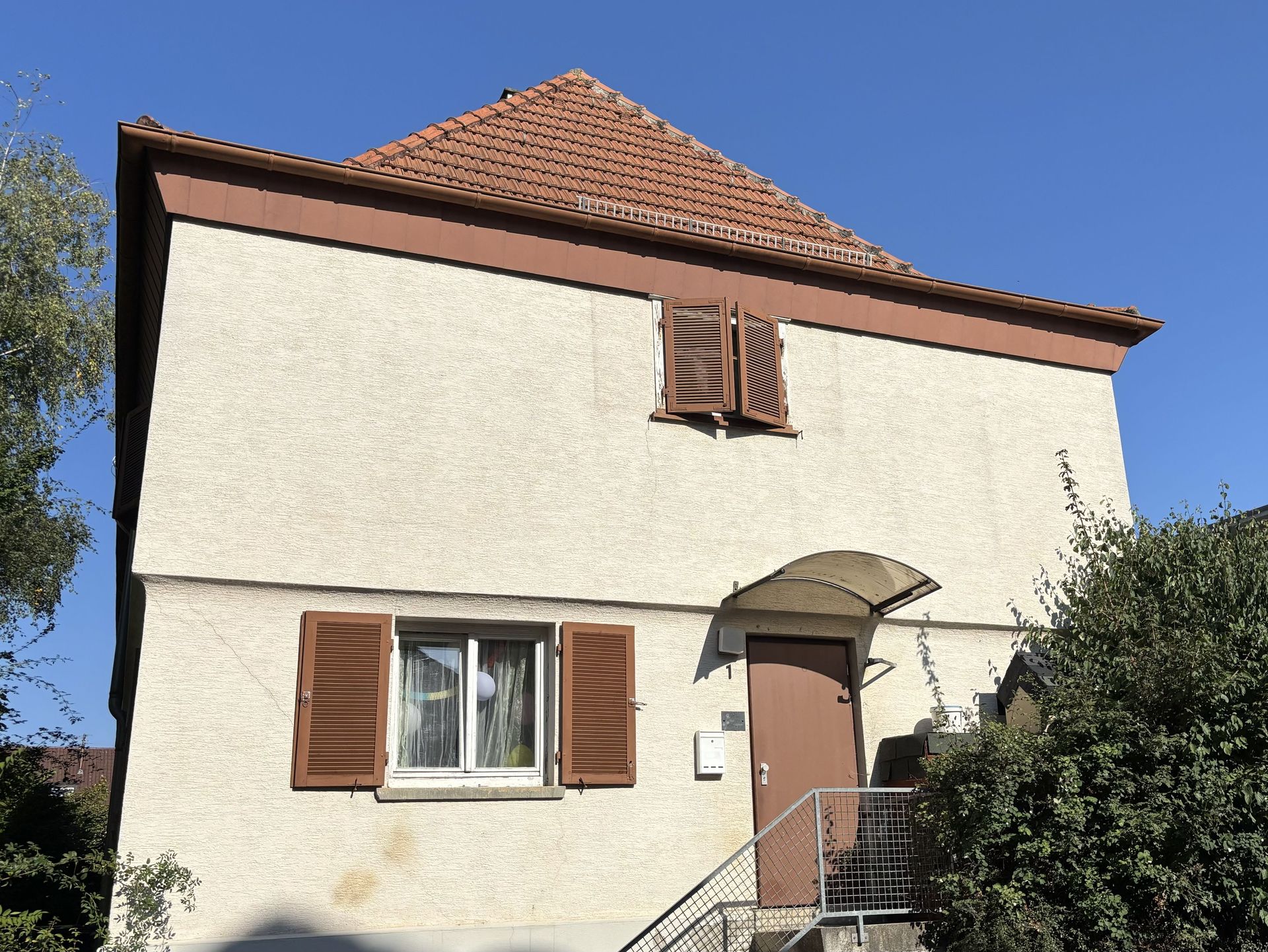 Das Bild zeigt ein kleines, helles Haus mit braunem Dach und braunen Fensterläden.  Eine Außentreppe führt zur Eingangstür, über der sich ein Vordach befindet.