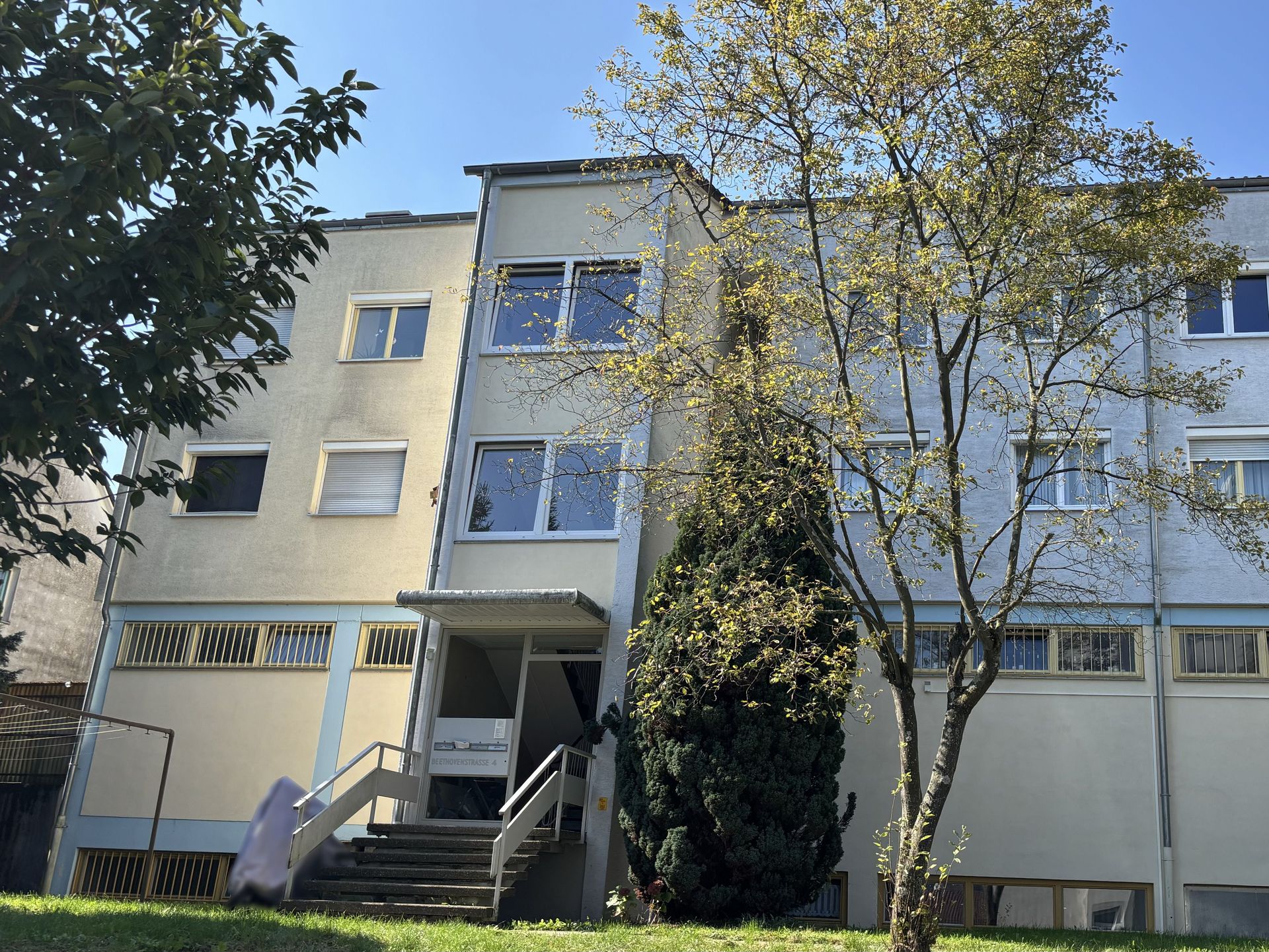 Das Bild zeigt ein Mehrfamilienhaus mit heller Fassade und mehreren Stockwerken. Vor dem Haus befindet sich eine Treppe, die zum Eingang führt und verschiedene Bäume.