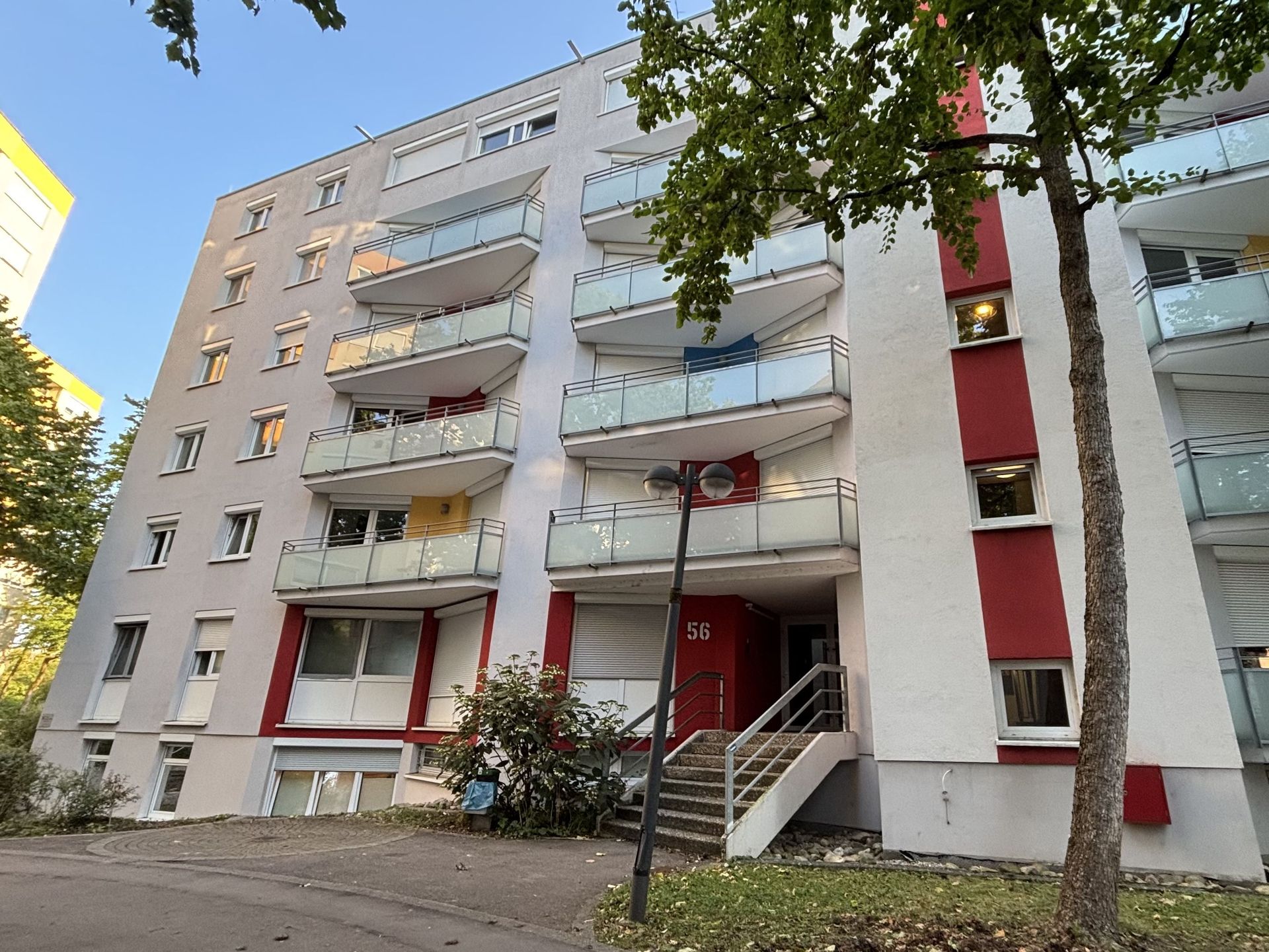 Das Bild zeigt ein Mehrfamilienhaus mit weißer Fassade, das durch rote Akzente an den Balkonen und dem Eingangsbereich aufgelockert wird.  Vor dem Haus befindet sich eine Treppe, die zum Eingang führt.
