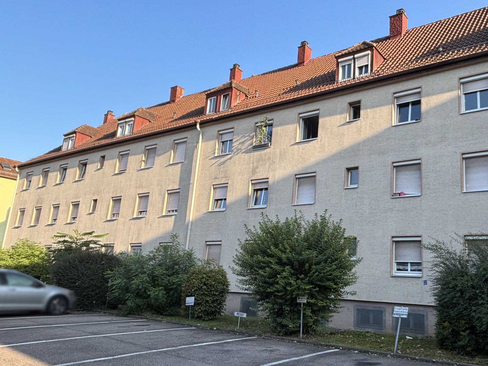 Das Bild zeigt ein Mehrfamilienhaus mit heller Fassade und rotem Dach. Vor dem Haus befindet sich ein Parkplatz mit einem geparkten Auto.