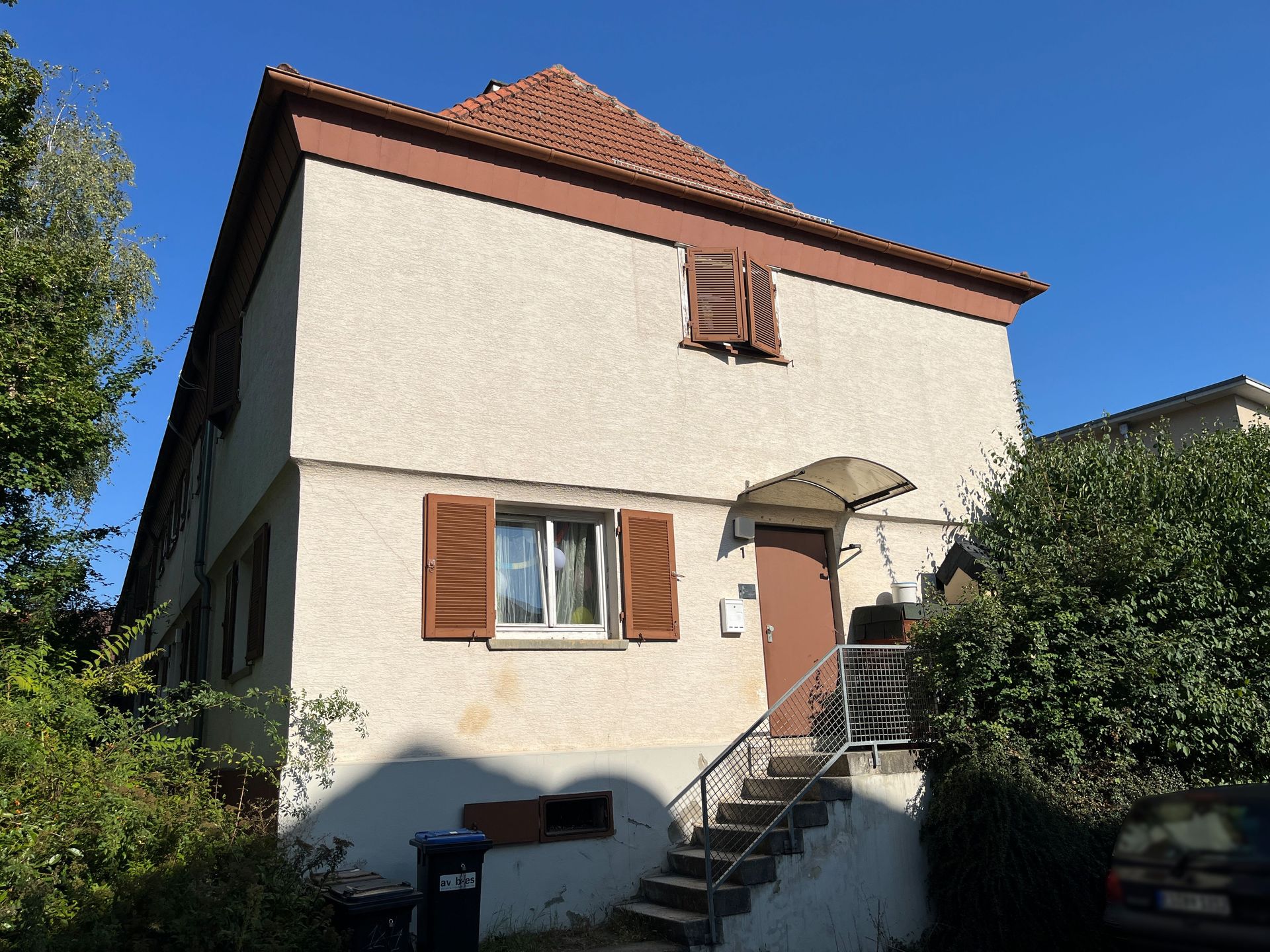 Das Bild zeigt ein kleines, hell verputztes Haus mit braunem Dach und braunen Fensterläden. Eine Außentreppe führt zum Eingang, der mit einem Vordach geschützt ist.