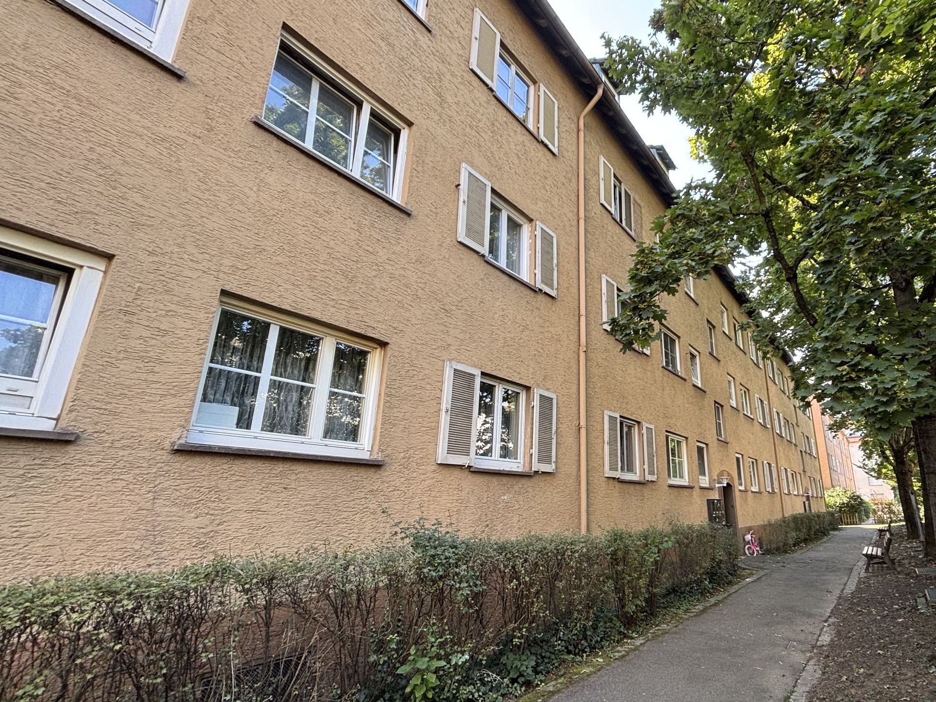  Das Bild zeigt ein Mehrfamilienhaus mit hellbrauner Fassade und weißen Fensterläden.  Ein schmaler Weg führt am Haus entlang und wird von einer Hecke und Bäumen gesäumt.