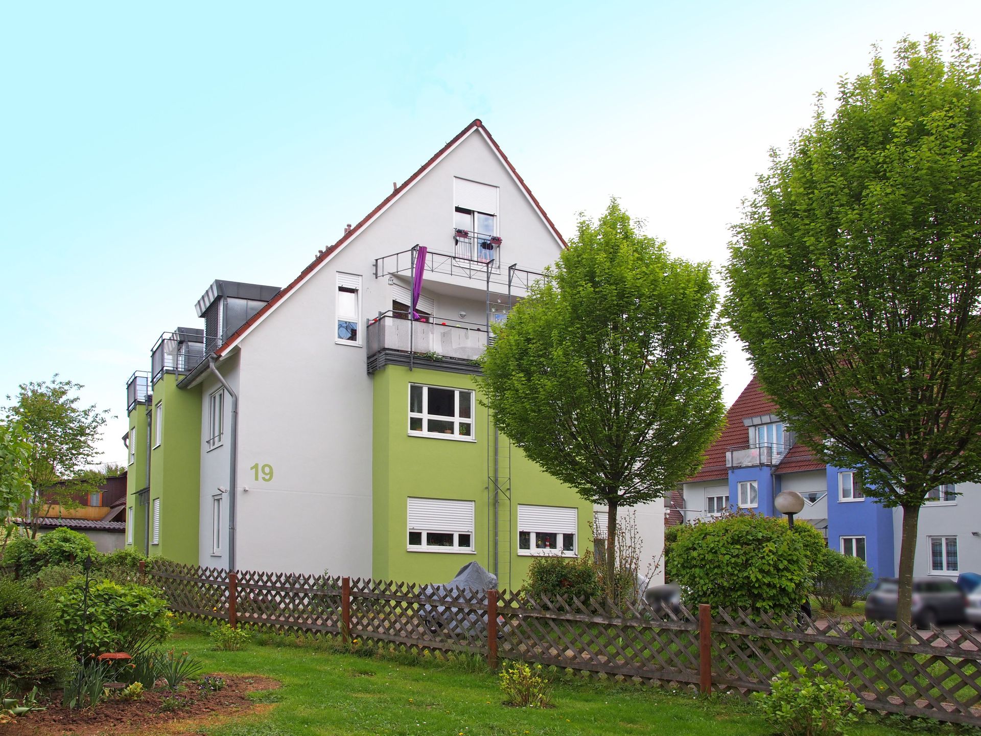 Das Bild zeigt ein Mehrfamilienhaus mit weißer und hellgrüner Fassade. Das Haus ist von einem Garten mit Bäumen und einem Holzzaun umgeben.