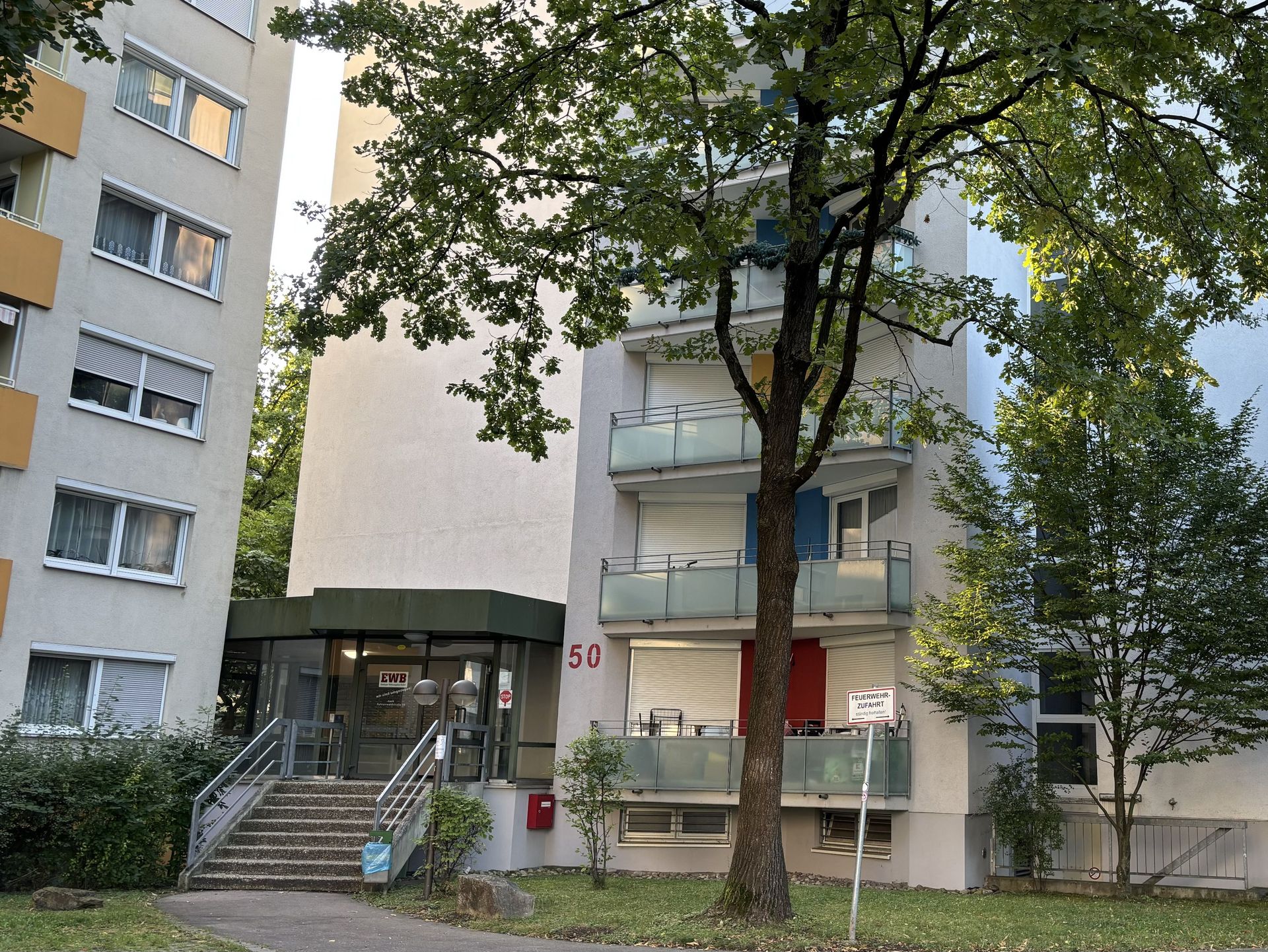 Das Bild zeigt ein mehrstöckiges Wohngebäude mit weißer Fassade und Balkonen, vor dem ein großer Baum steht.  Eine Treppe führt zum Eingangsbereich, der durch einen verglasten Anbau gekennzeichnet ist.
