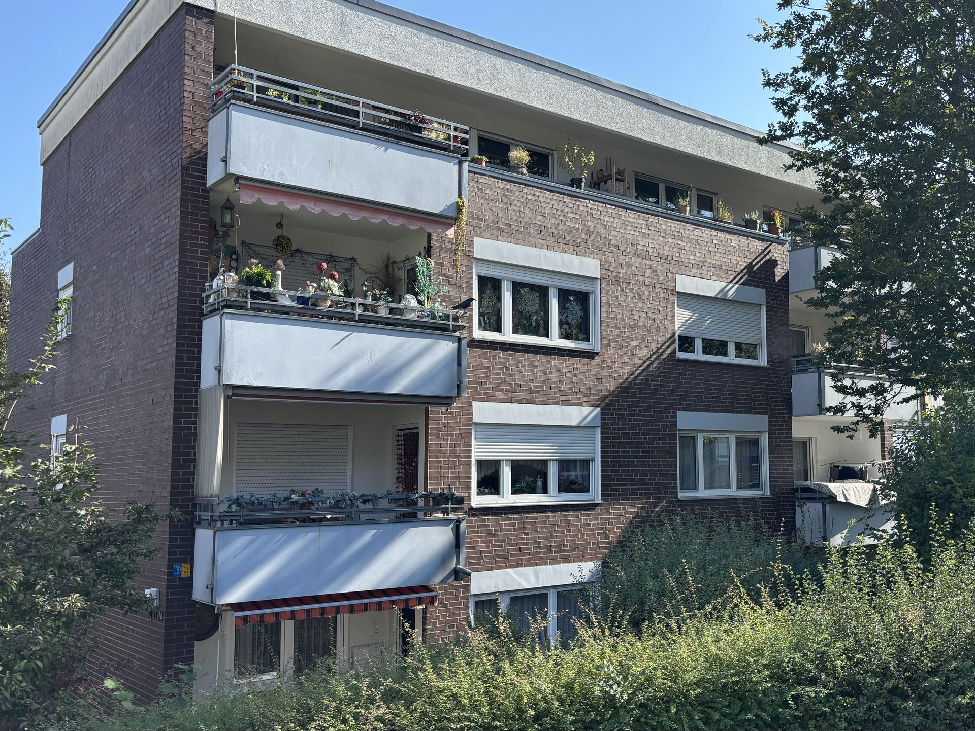 Das Bild zeigt ein Mehrfamilienhaus mit brauner Klinkerfassade und weißen Balkonen. Das Haus ist von Sträuchern und Bäumen umgeben.