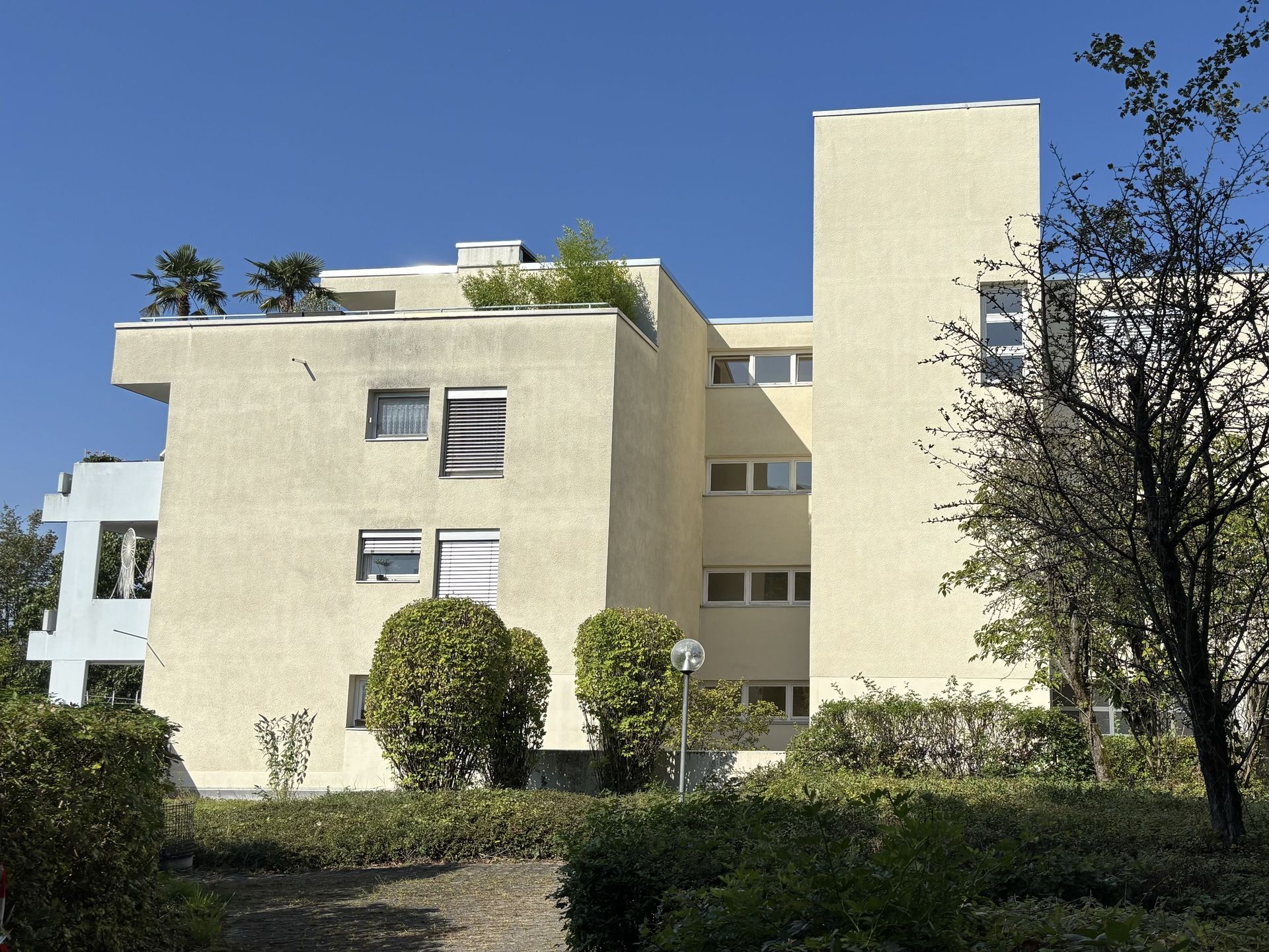 Das Bild zeigt ein Mehrfamilienhaus mit heller Fassade und einem zurückgesetzten Dachgeschoss. Vor dem Haus befindet sich eine Grünfläche mit Büschen und Bäumen.