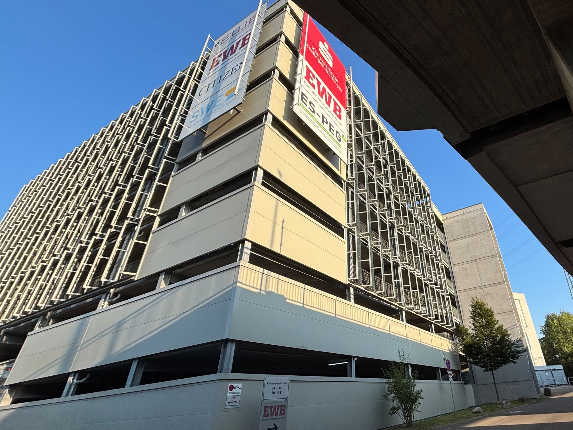 Das Bild zeigt ein mehrstöckiges Parkhaus mit einer Fassade aus hellen Betonplatten und einer modernen, gitterartigen Verkleidung.  An der Seite des Gebäudes sind mehrere Banner mit dem Logo der EWB und weiteren Schriftzügen angebracht.