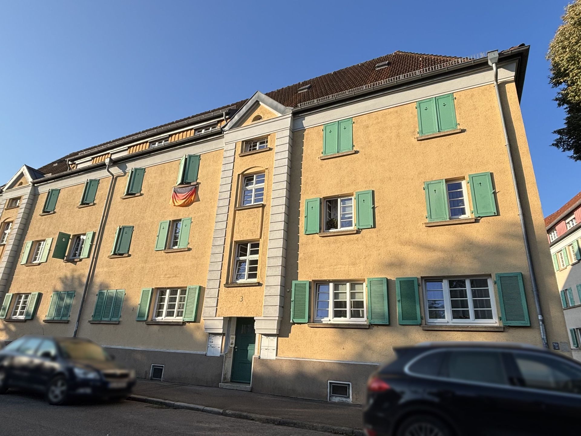 Das Bild zeigt ein Mehrfamilienhaus mit hellgelber Fassade und grünen Fensterläden. An der Fassade hängt eine Deutschlandflagge und vor dem Haus stehen zwei Autos am Straßenrand.