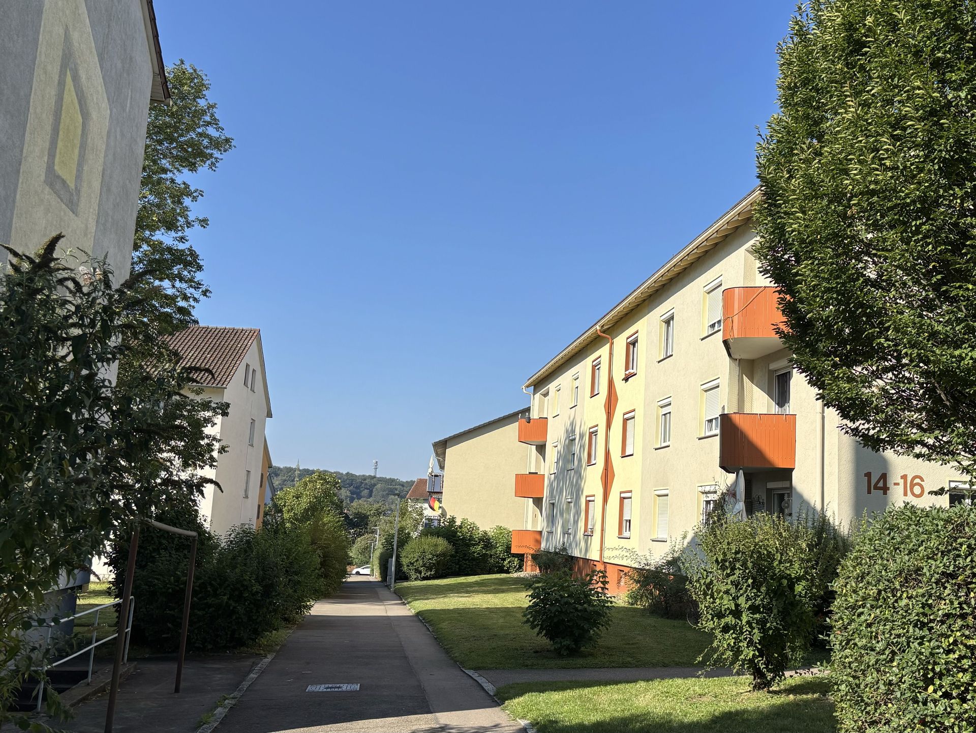 Das Bild zeigt eine Wohnsiedlung mit Mehrfamilienhäusern und einem Fußweg. Die Häuser haben helle Fassaden und teilweise orangefarbene Balkone.