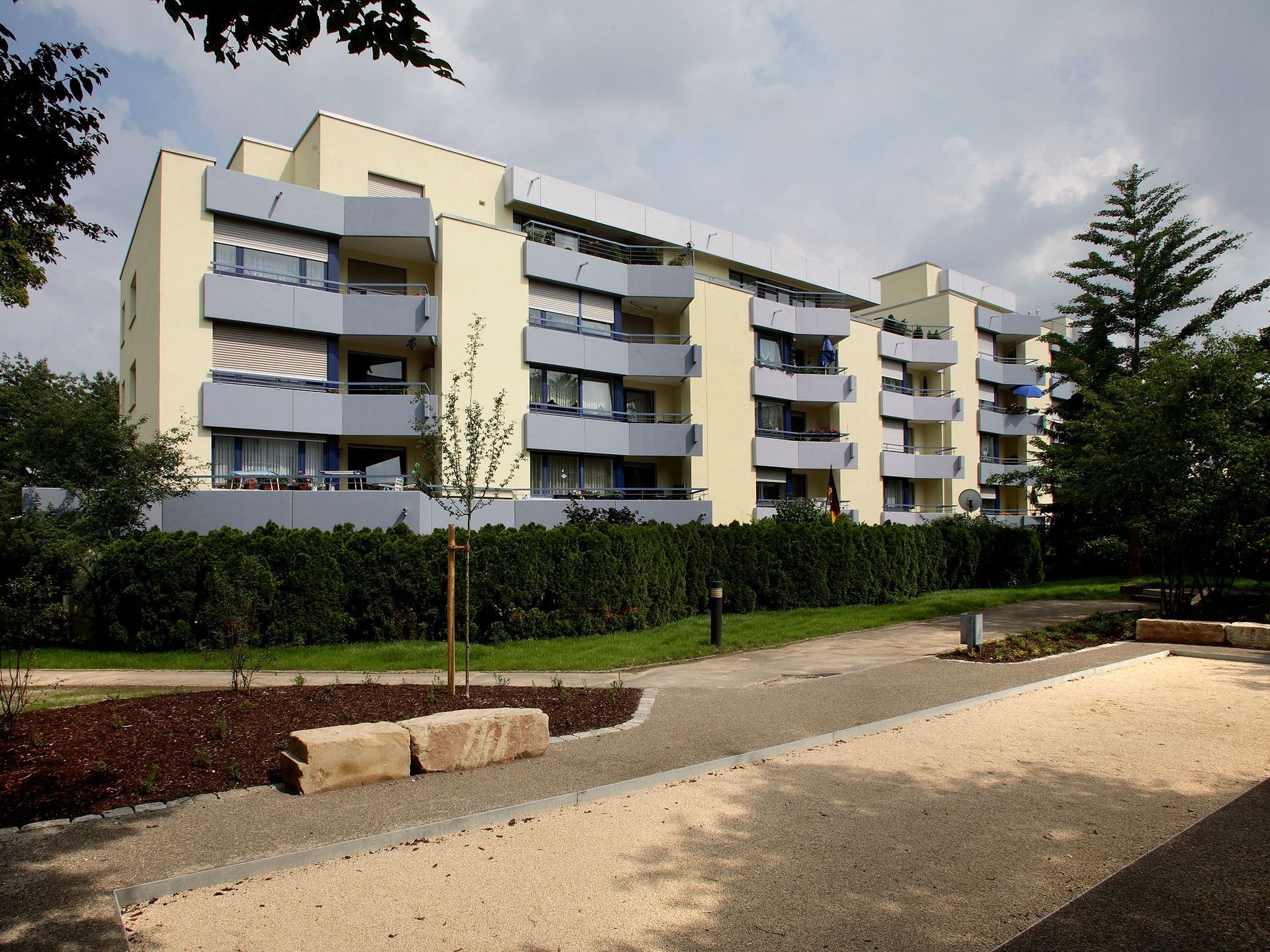 Das moderne Mehrfamilienhaus besticht durch seine hellgelbe Fassade, graue Balkone und zahlreiche Wohneinheiten, die sich über mehrere Stockwerke erstrecken. Es ist umgeben von Rasenflächen, Bäumen und Sträuchern, gepflasterten Wegen und einem Sandkasten.