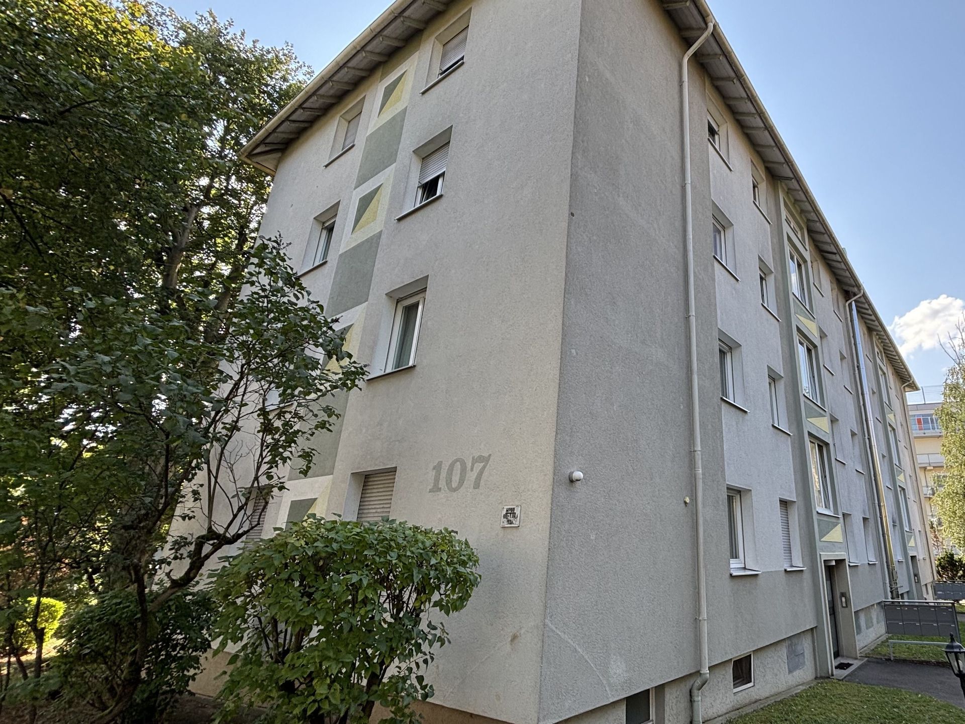 Das Bild zeigt ein Mehrfamilienhaus mit grauer Fassade und gelben und grünen Akzenten um die Fenster. Vor dem Haus stehen einige Bäume und Büsche, die das Gebäude teilweise verdecken.
