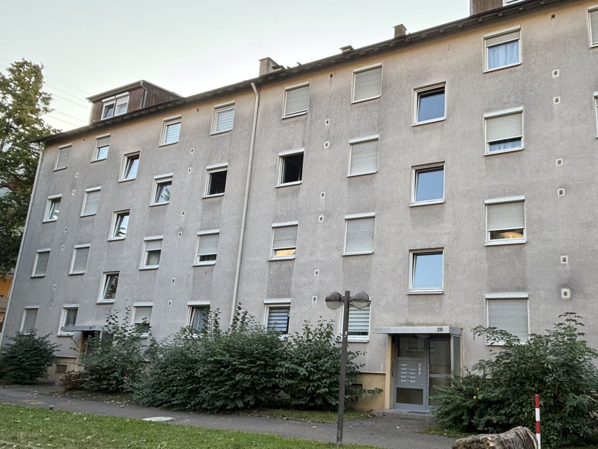 Das Bild zeigt ein schlichtes, graues Mehrfamilienhaus mit mehreren Stockwerken und zahlreichen Fenstern, die mit Rollläden ausgestattet sind. Vor dem Haus befindet sich ein gepflasterter Gehweg und eine grüne Hecke.