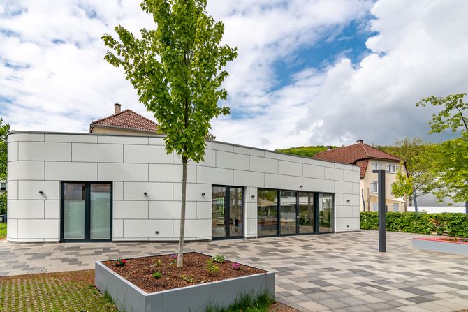 Das Bild zeigt ein Gebäude mit heller Fassade und einem Baum.