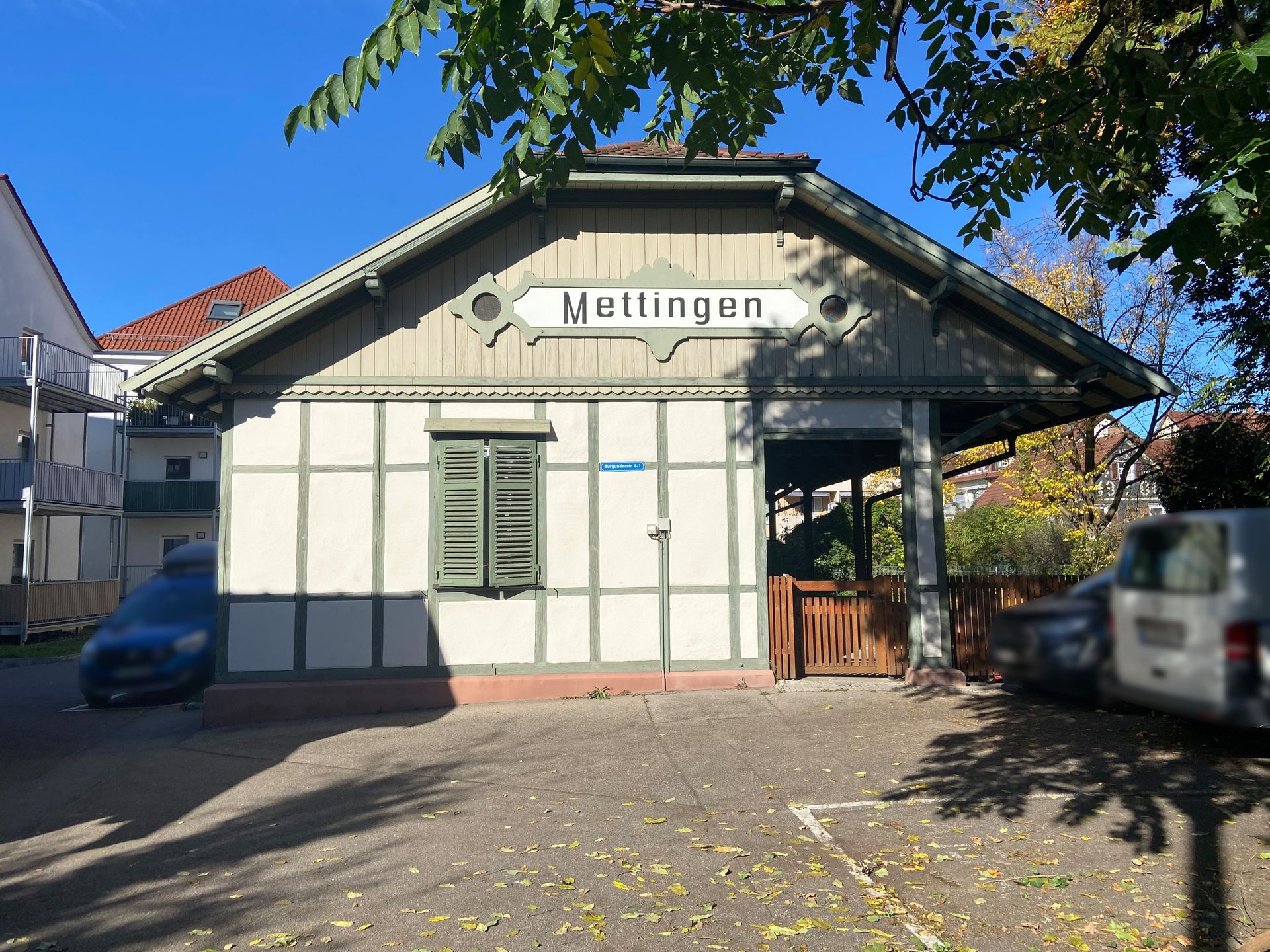 Das Bild zeigt ein kleines Gebäude mit der Aufschrift "Mettingen" und grünen Fensterläden. Das Gebäude hat eine Holzfassade mit Fachwerk-Optik und ein Satteldach.