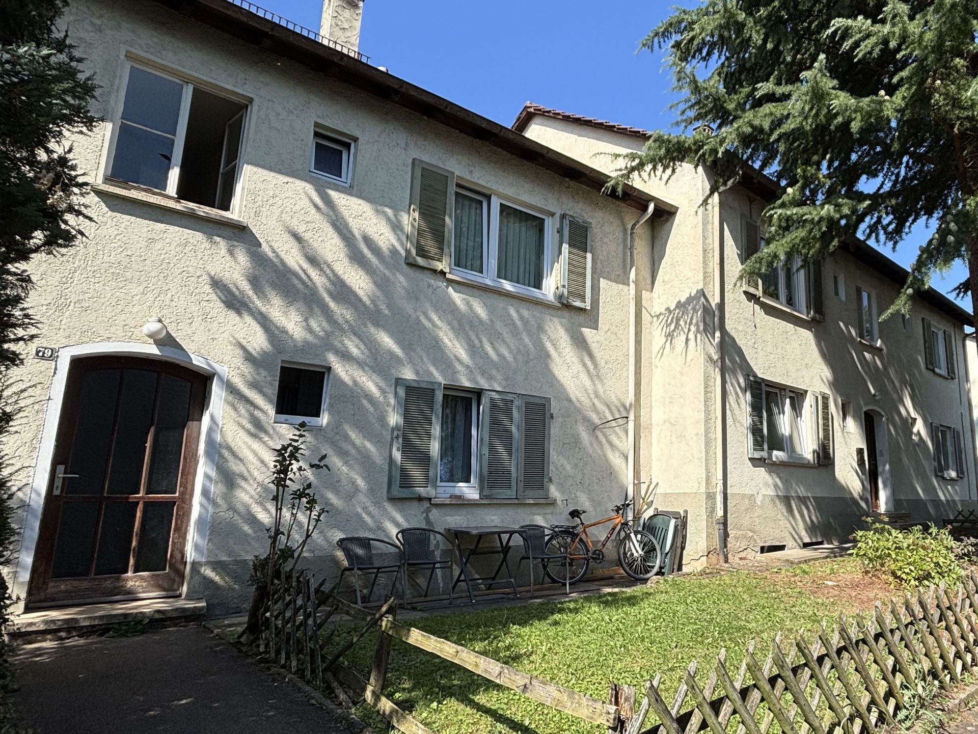Das Bild zeigt zwei Mehrfamilienhäuser mit heller Fassade und dunklen Dächern. Vor den Häusern ist eine Rasenfläche umzäunt von einem braunen Holzzaun. Dort stehen Sitzmöglichkeiten und ein Fahrrad.