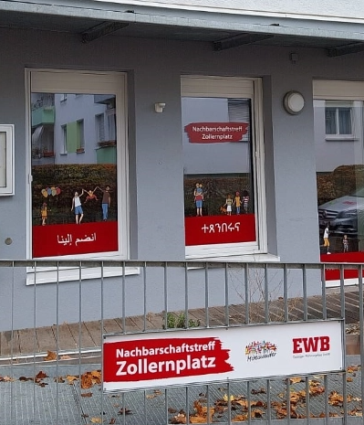 Das Bild zeigt eine Hausfront mit Fenstern, die mit Gestaltungselementen beklebt sind. Vorne im Bild sieht man ein Geländer und dort ist ein Schild befestigt. Link zum Bereich Nachbarschaftstreff Zollernplatz.