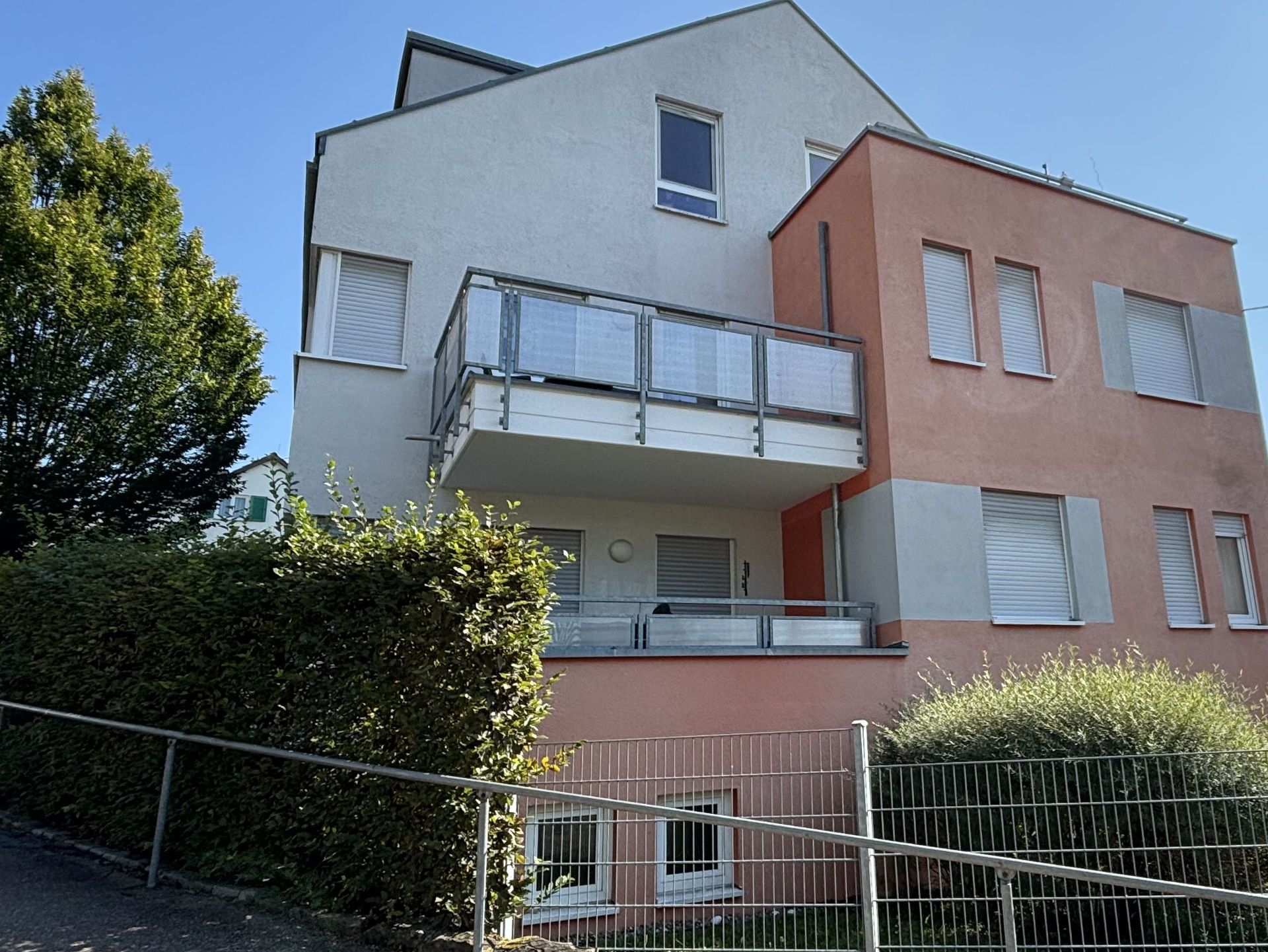 Das Bild zeigt ein modernes Mehrfamilienhaus mit zwei Balkonen. Die Fassade des Gebäudes ist in zwei Farben gestaltet: Weiß und einem hellen Rotton.