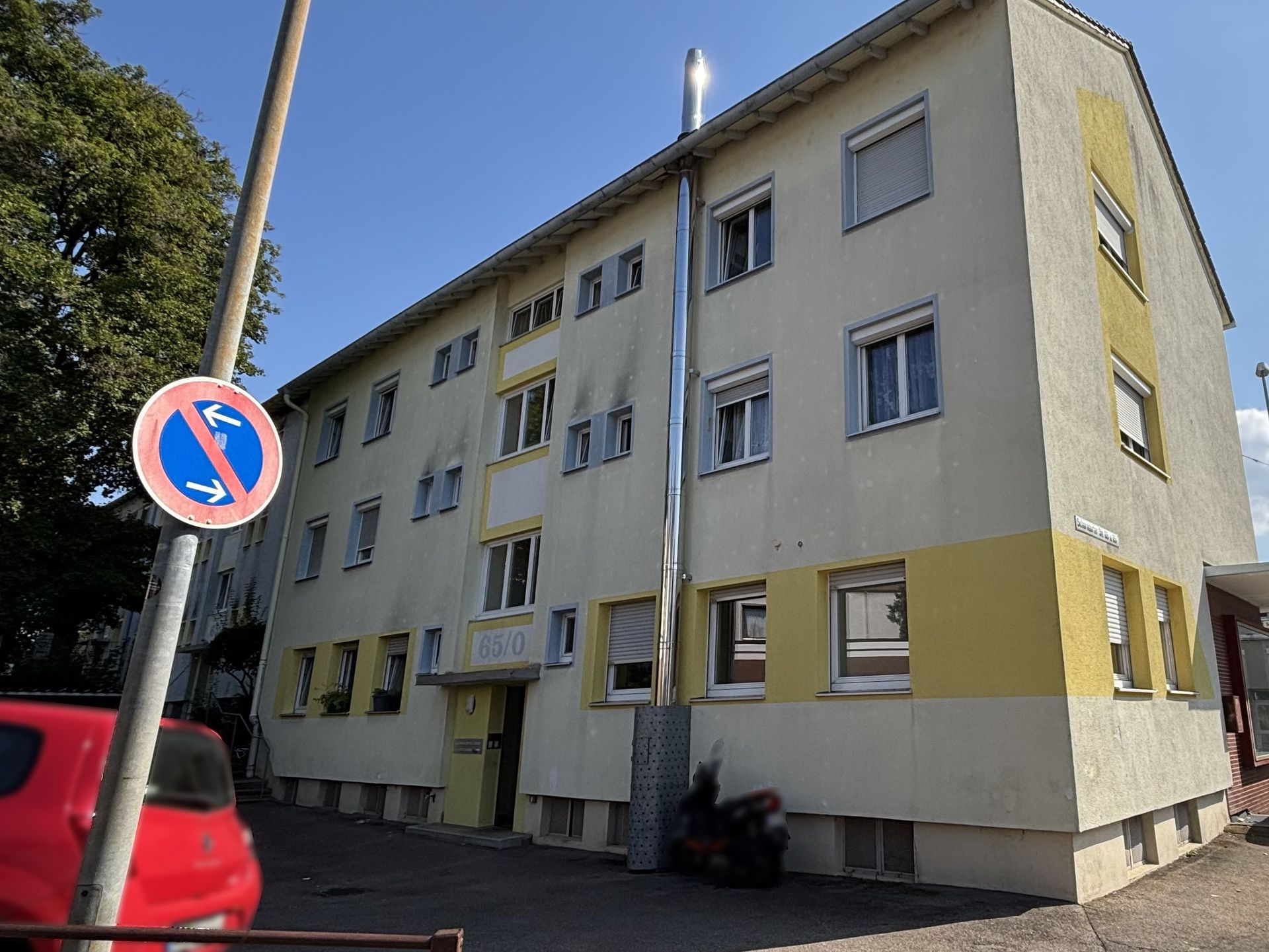 Das Bild zeigt ein mehrstöckiges Mehrfamilienhaus mit grauer und gelber Fassade. Zwischen den Fenstern ist eine silberne Röhre angebracht. Vor dem Haus verläuft ein gepflasteter Weg.