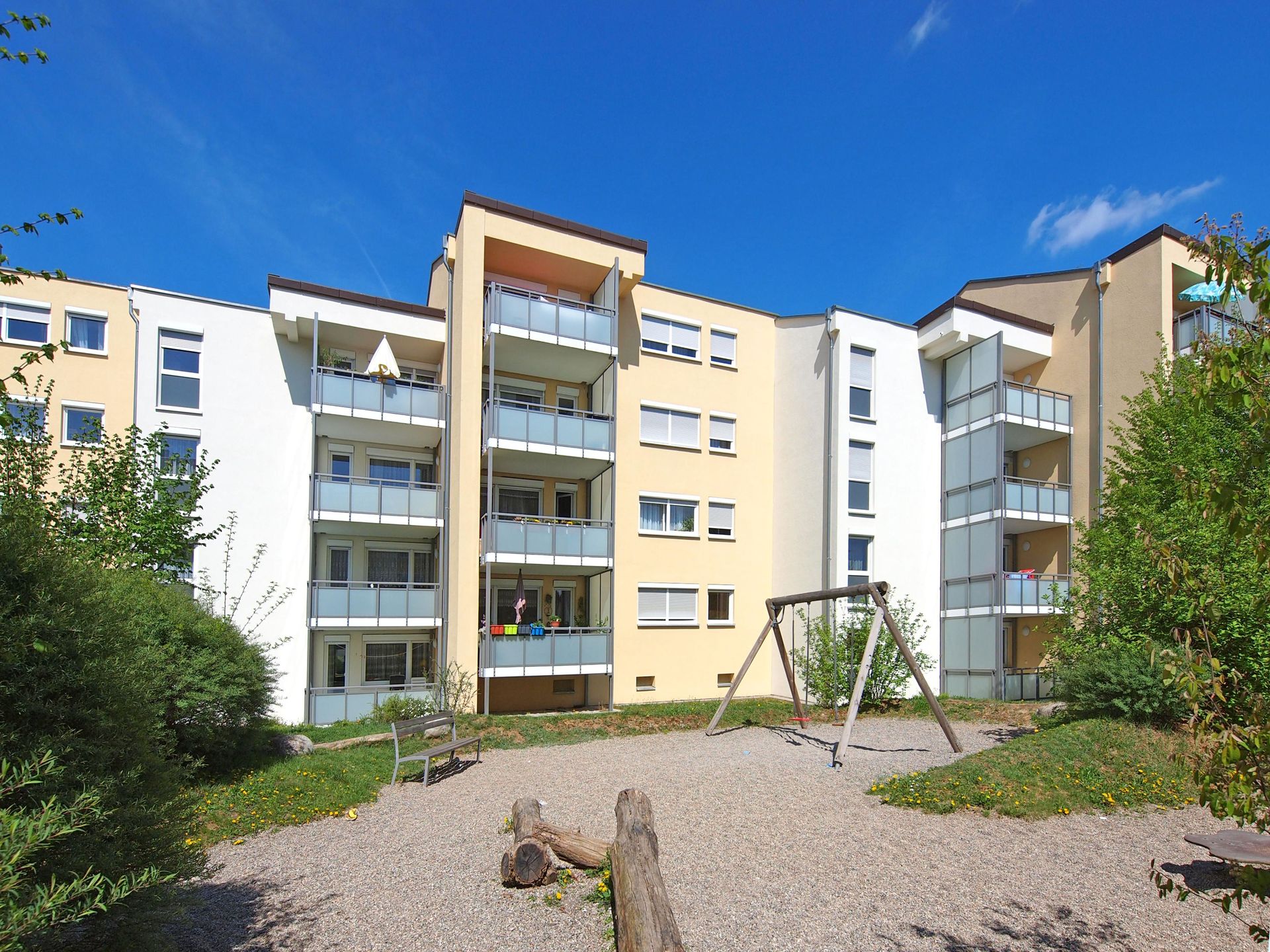 Das Bild zeigt ein Mehrfamilienhaus mit Balkonen. Vor dem Haus befindet sich ein kleiner Spielplatz mit Schaukel und Sitzgelegenheiten.