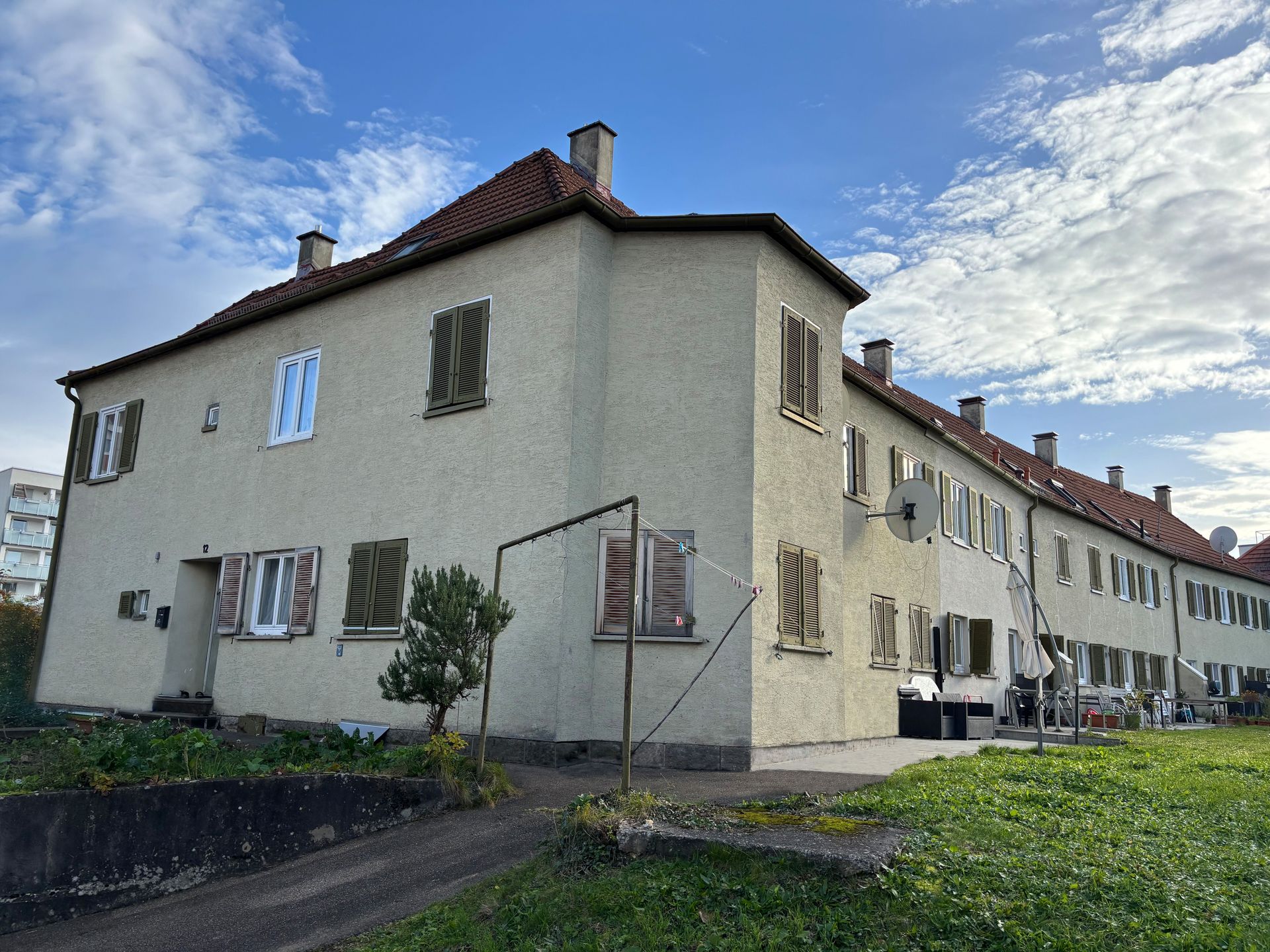 Das Bild zeigt ein Haus mit heller Fassade, braunen Fensterläden und dunklem Dach. Das Haus ist umgeben von Grünflächen.