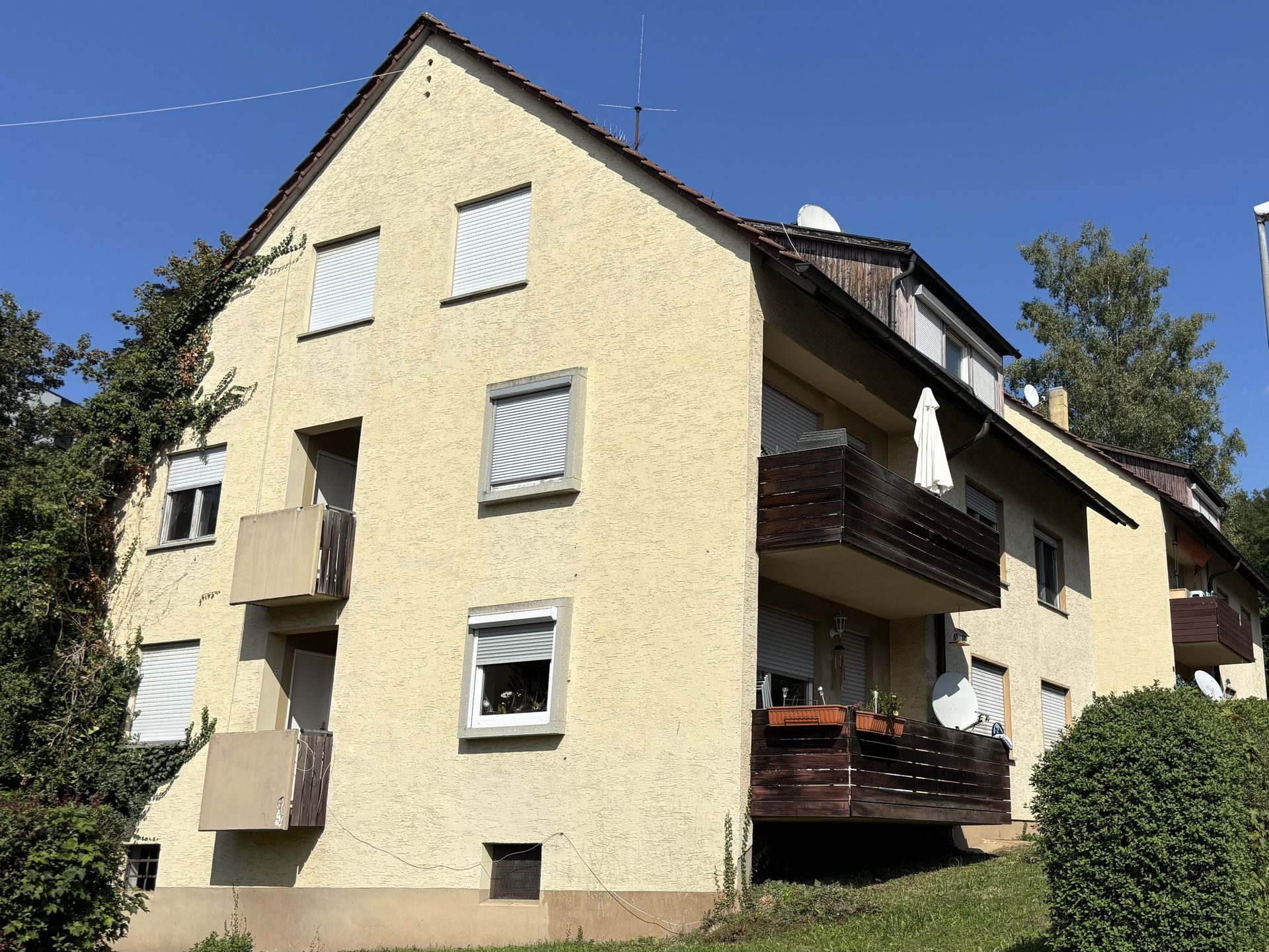 Das Bild zeigt ein Mehrfamilienhaus mit hellgelber Fassade, das über mehrere Balkone verfügt. Die Balkone haben dunkle Holzgeländer und sind teilweise mit Sonnenschirmen und Pflanzen ausgestattet.