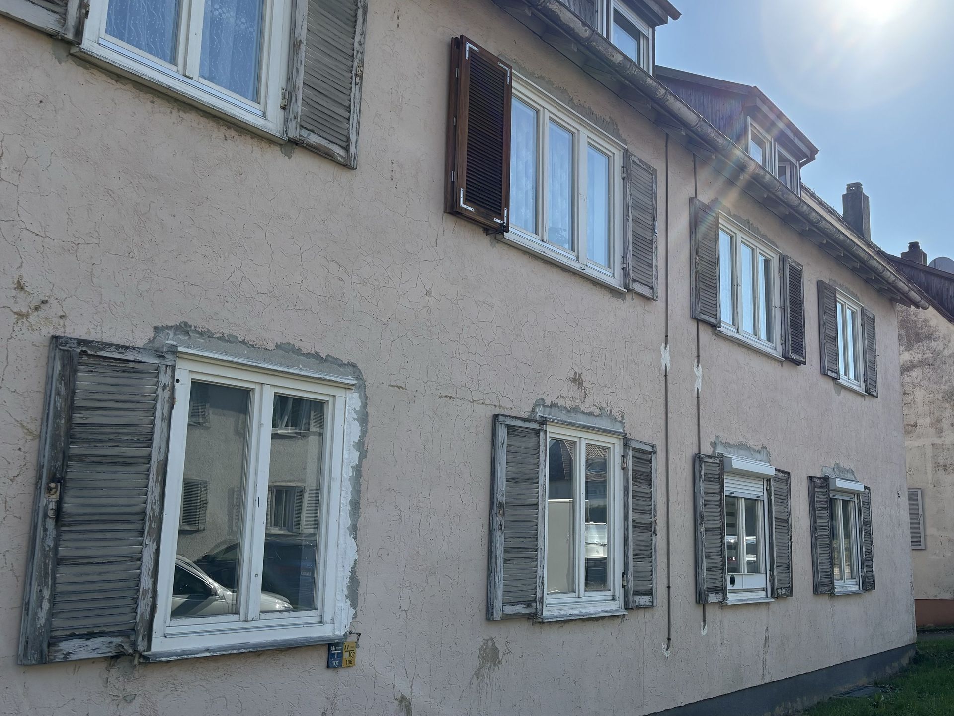 Das Bild zeigt ein Mehrfamilienhaus mit heller Fassade und Fenstern, die mit verschiedenen Fensterläden ausgestattet sind. An einigen Stellen blättert der Putz von der Fassade ab.